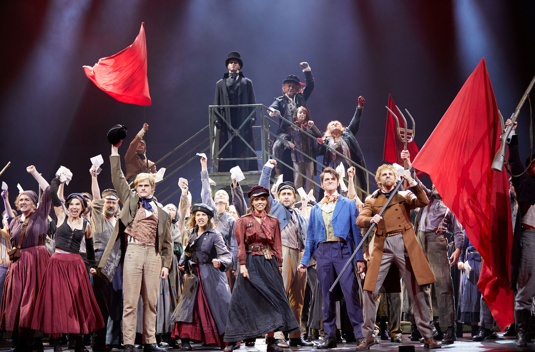 Marius und Enjolras stehen in der Menge des Ensembles. Es werden die Arme nach oben gestreckt und rote Fahnen wehen. Auf einem Podest hinter der Menge steht ein Mann in schwarzem Anzug mit Zylinder.