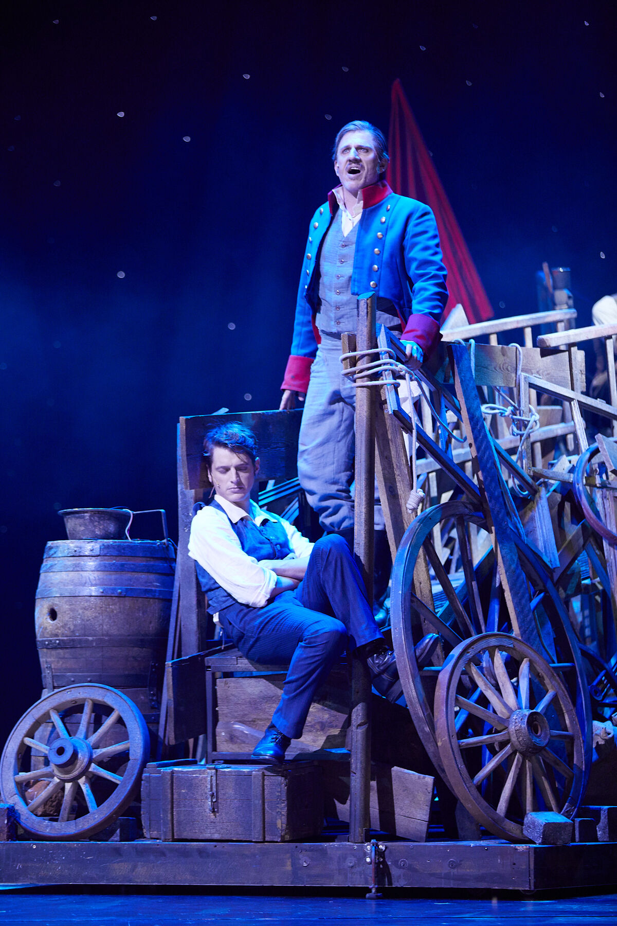 Marius sitzt auf Holzkisten und hat die Augen zu. um ihn herum stehen Kutschräder und Holzfässer. Hinter ihm steht Jean Valjean und singt. Der Hintergrund ist blau.
