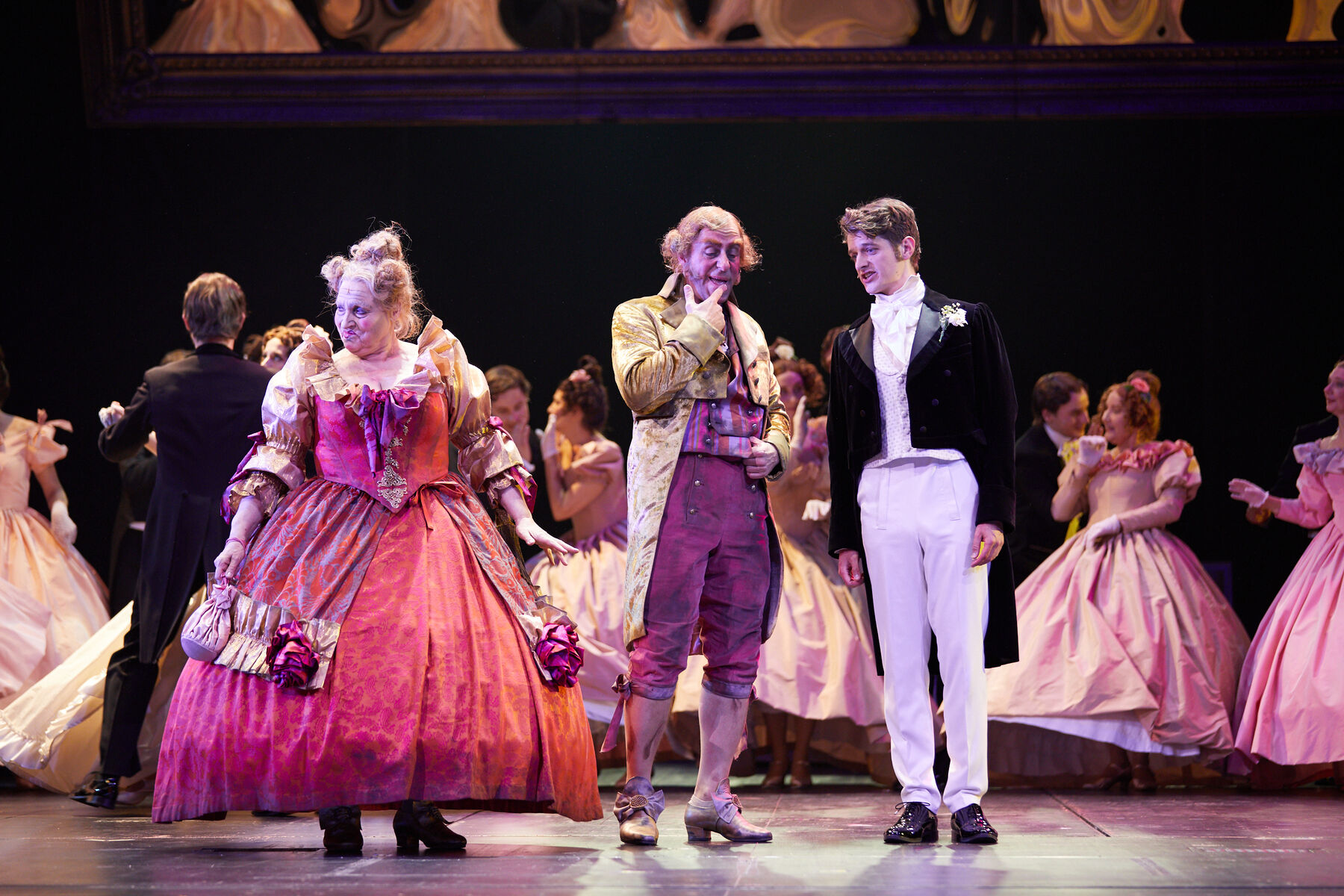 Madame Thénardier in einem pinken Kleid, Thénardier und Marius stehen nebeneinander auf der Bühne. Die Männer unterhalten sich. Im Hintergrund sieht man Frauen in rosa Kleidern mit Männern tanzen.