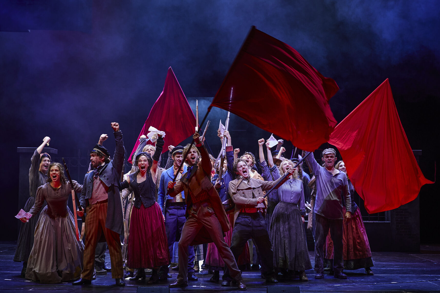 Marius und Enjolras stehen in der Menge des Ensembles. Alle haben die arme nach oben gestreckt und schreien. Drei rote Fahnen wehen.