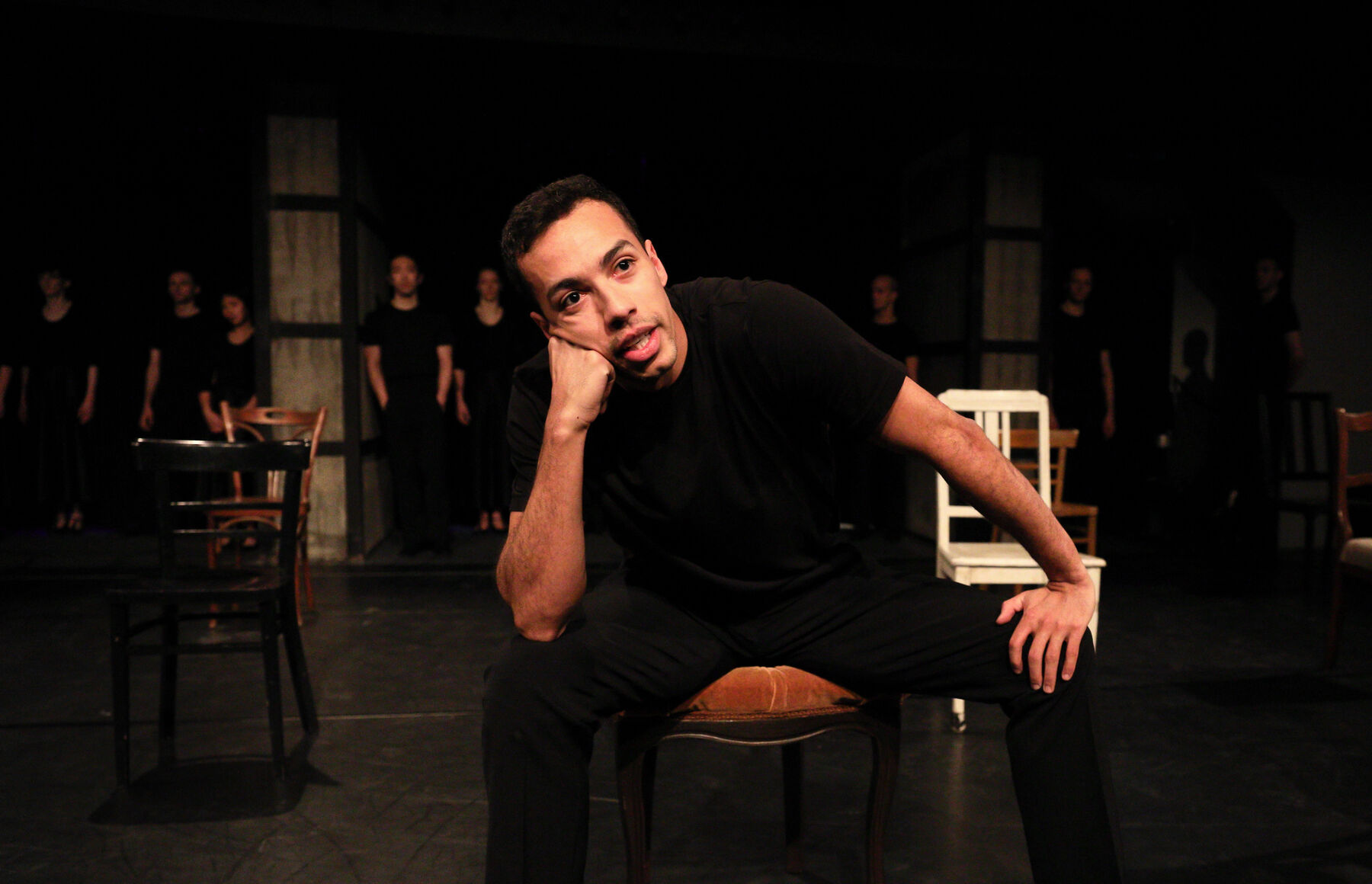 A man is sitting on a chair. Chairs are standing behind him. In the background can be seen other dancers standing in a line.