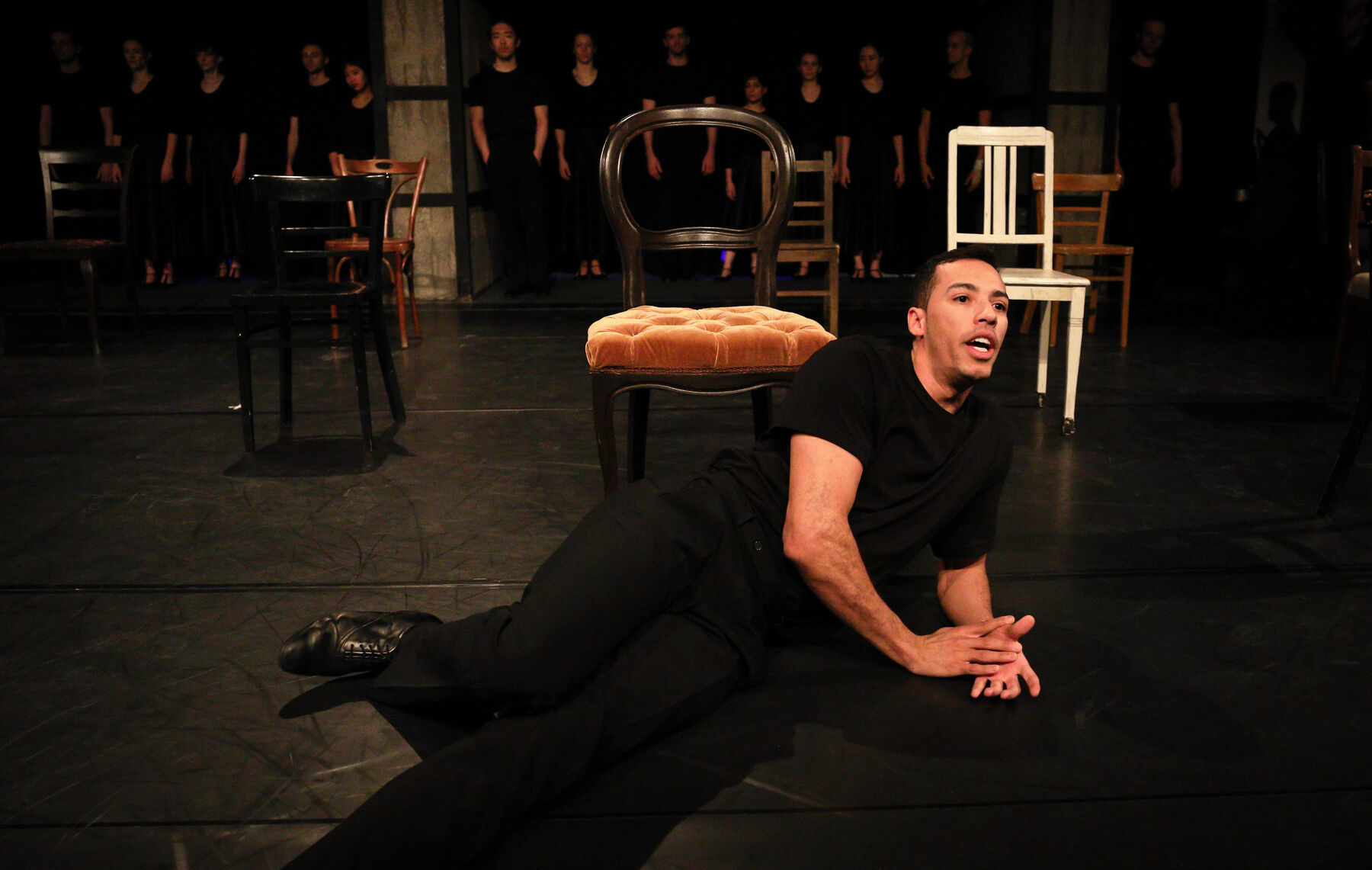 A Dancer is lying on the floor. Chairs are standing behind him. In the background other dancers, standing in a line, can be seen.