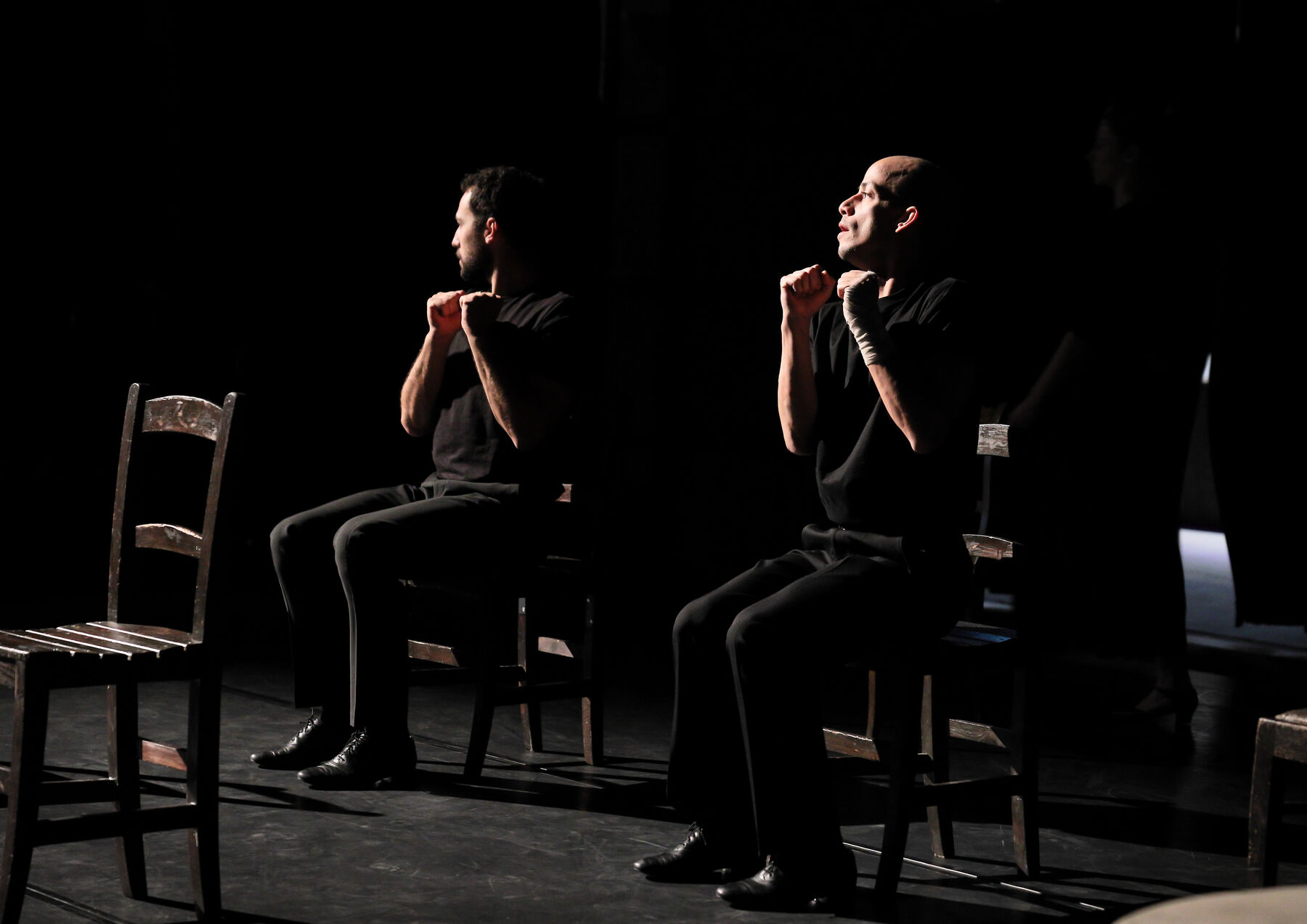 dancers, dressed in black, are sitting on chairs, in a dark room.
