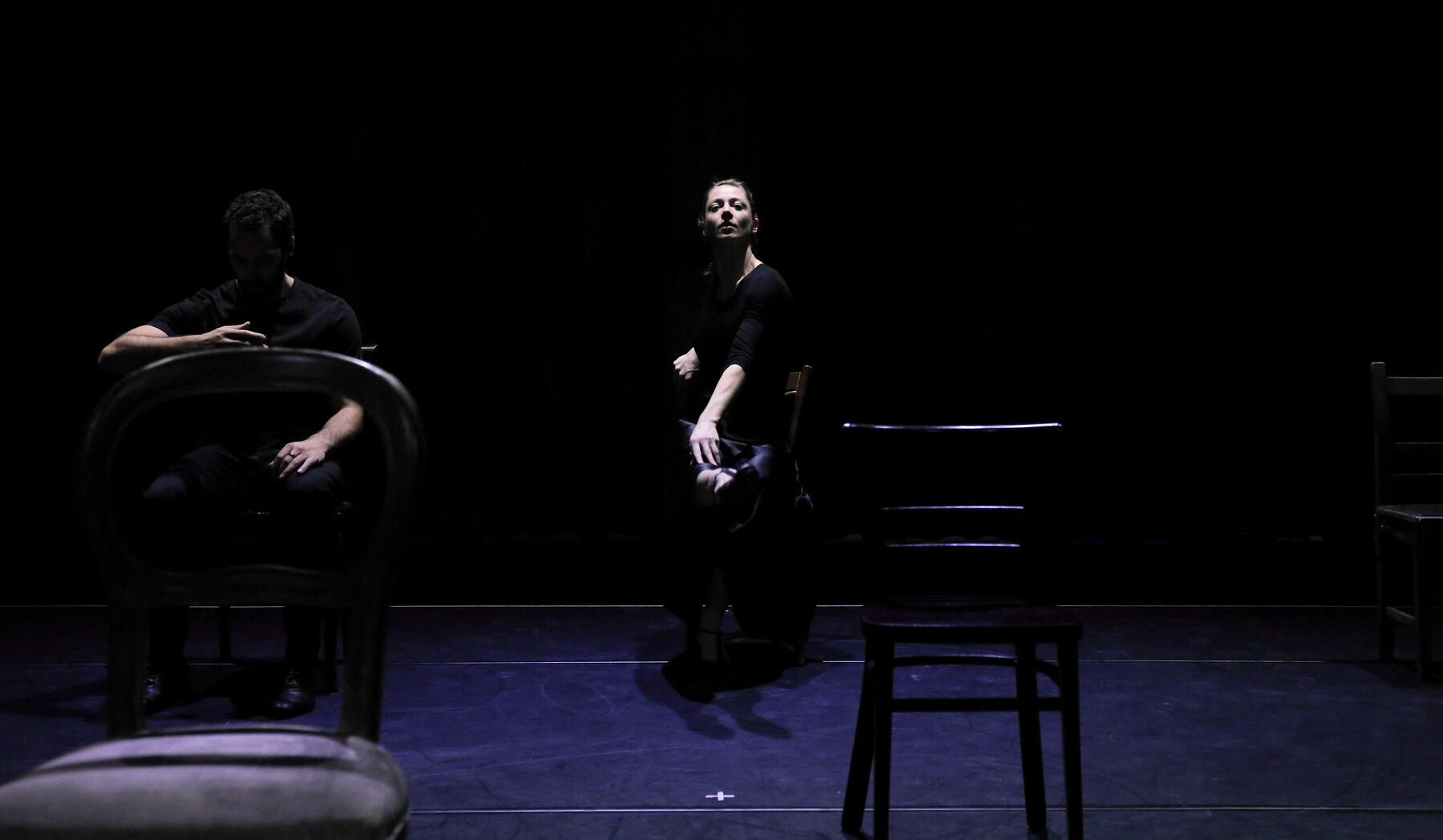 Dancers are sitting on chairs, in a dark room.