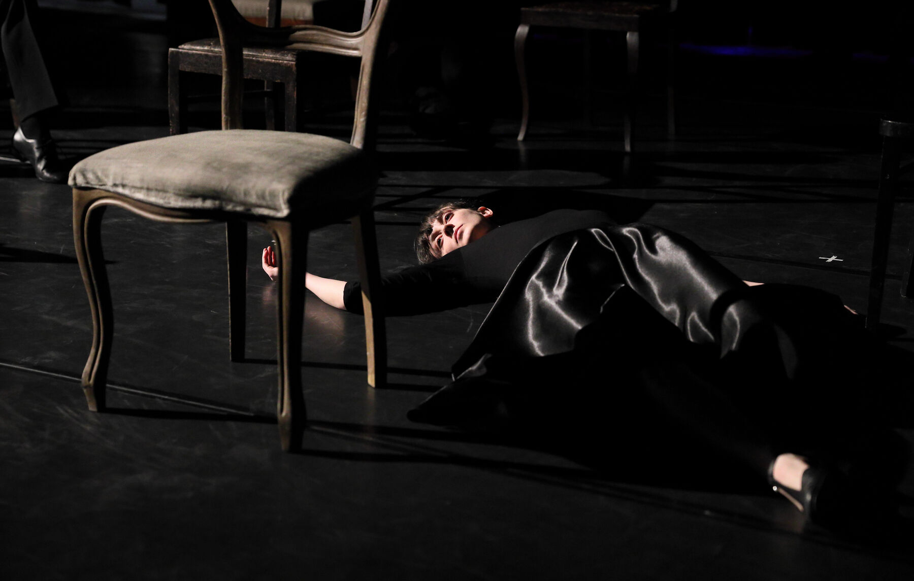 A woman, dressed in black, is lying on the floor. Chairs can be seen, also.
