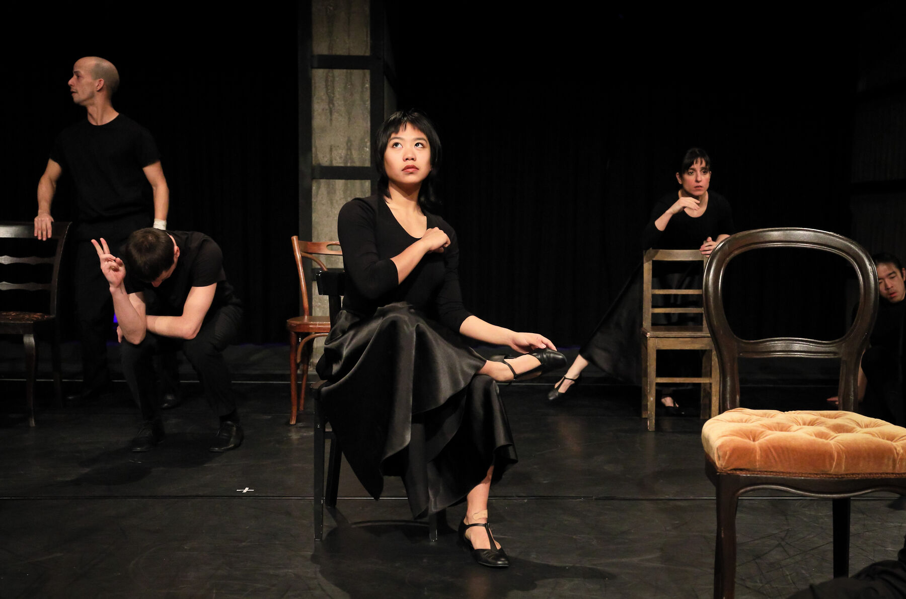 In the middle, a woman is sitting on a chair. Other chairs and dancers can be seen in the room.