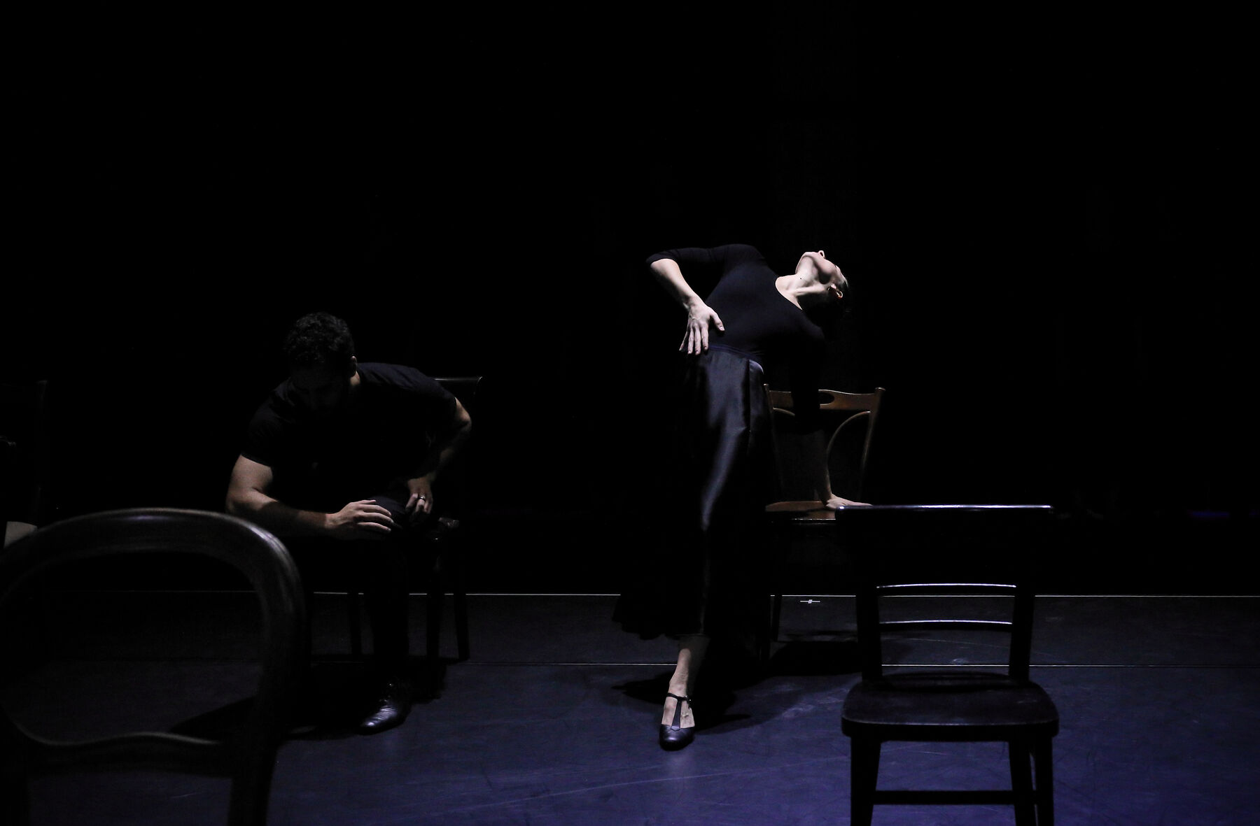Chairs are standing in a dark room. A man is sitting on one of the chairs, a woman is dancing by another.