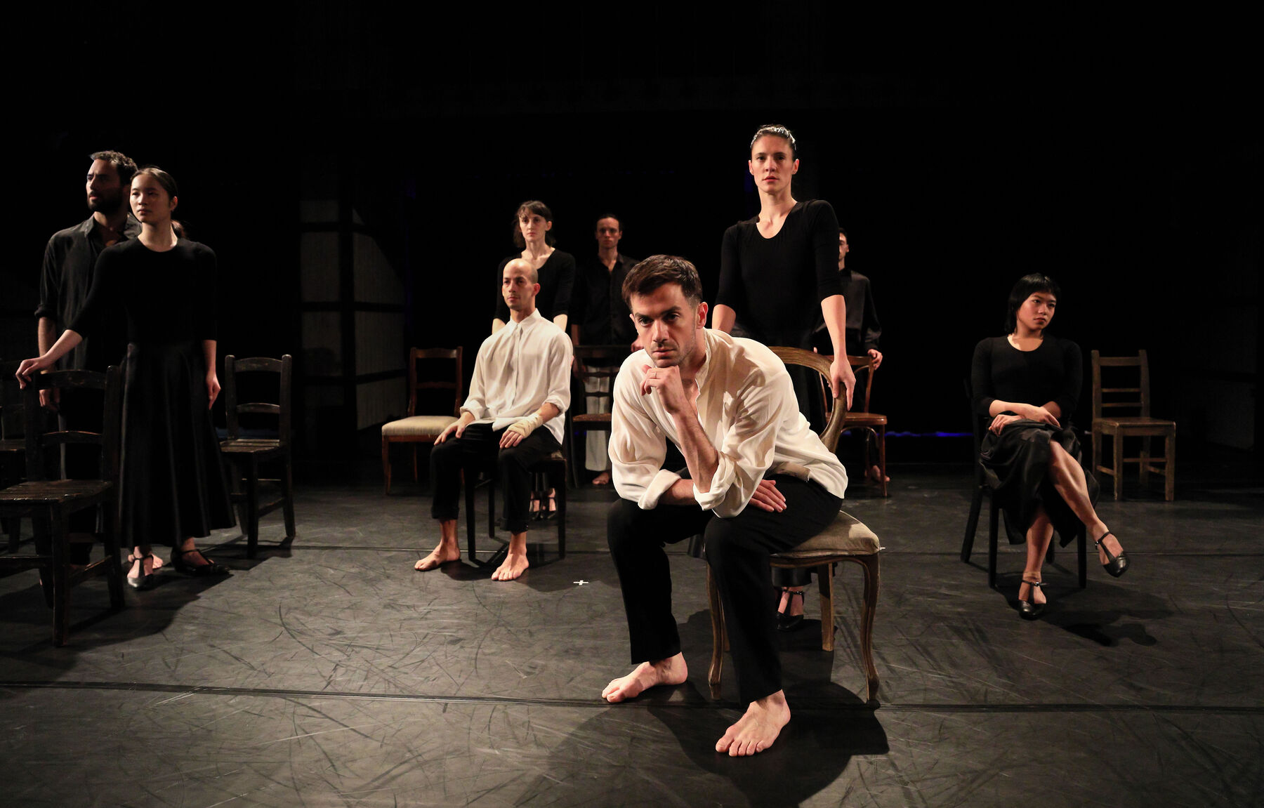 Chairs are standing in a dark room.  Dancers are sitting on the chairs, or standing behind.