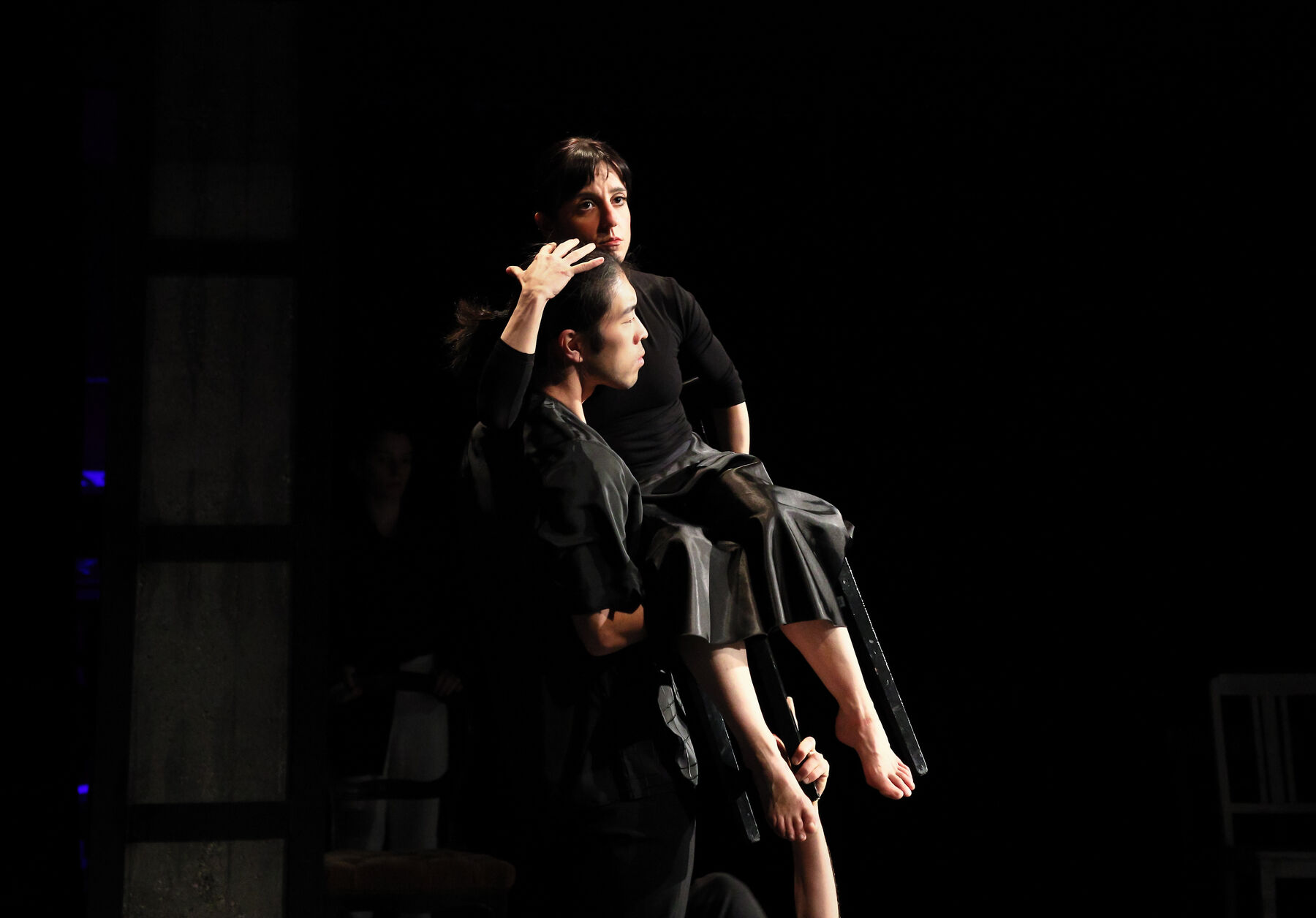 two dancers, dressed in black, can be seen. the woman is sitting on a chair, the man is holding her.