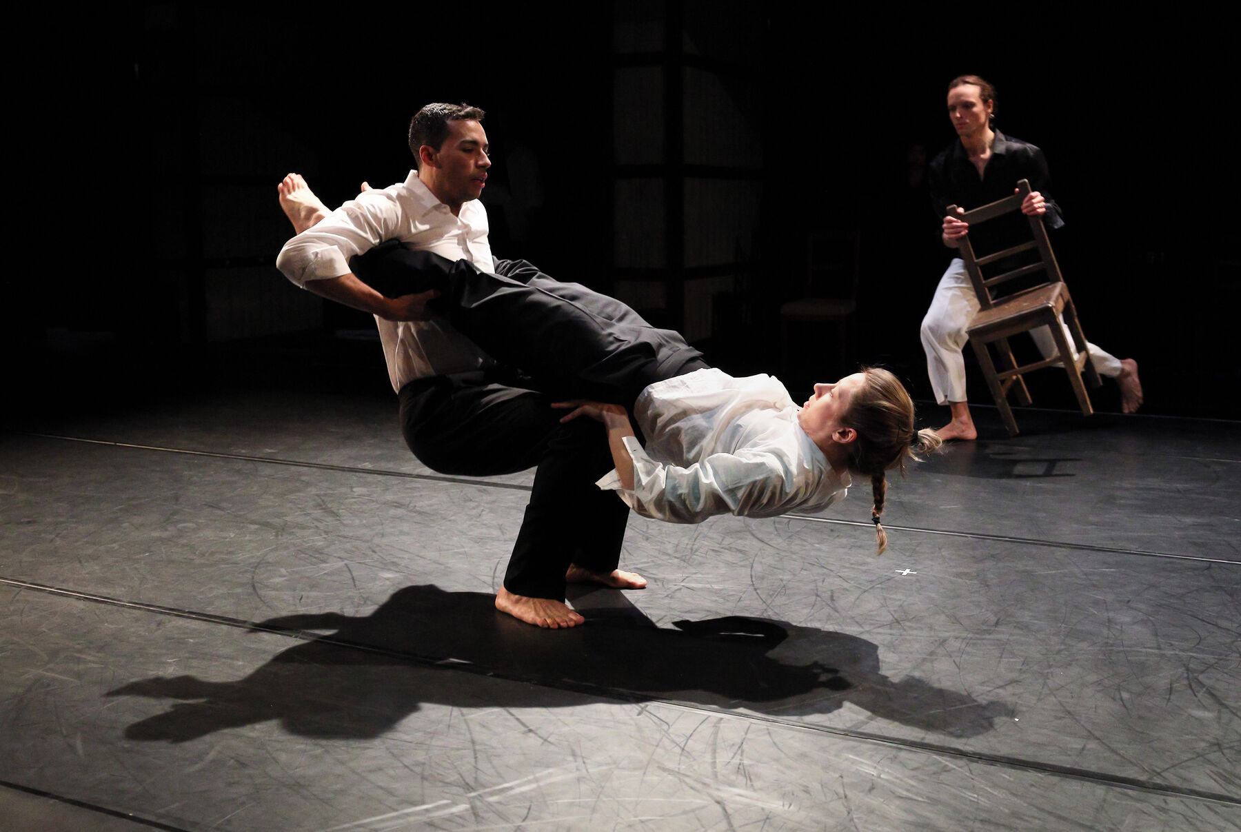 In the foreground, can be seen two dancers. In the back, can be seen another dancer, with a chair.
