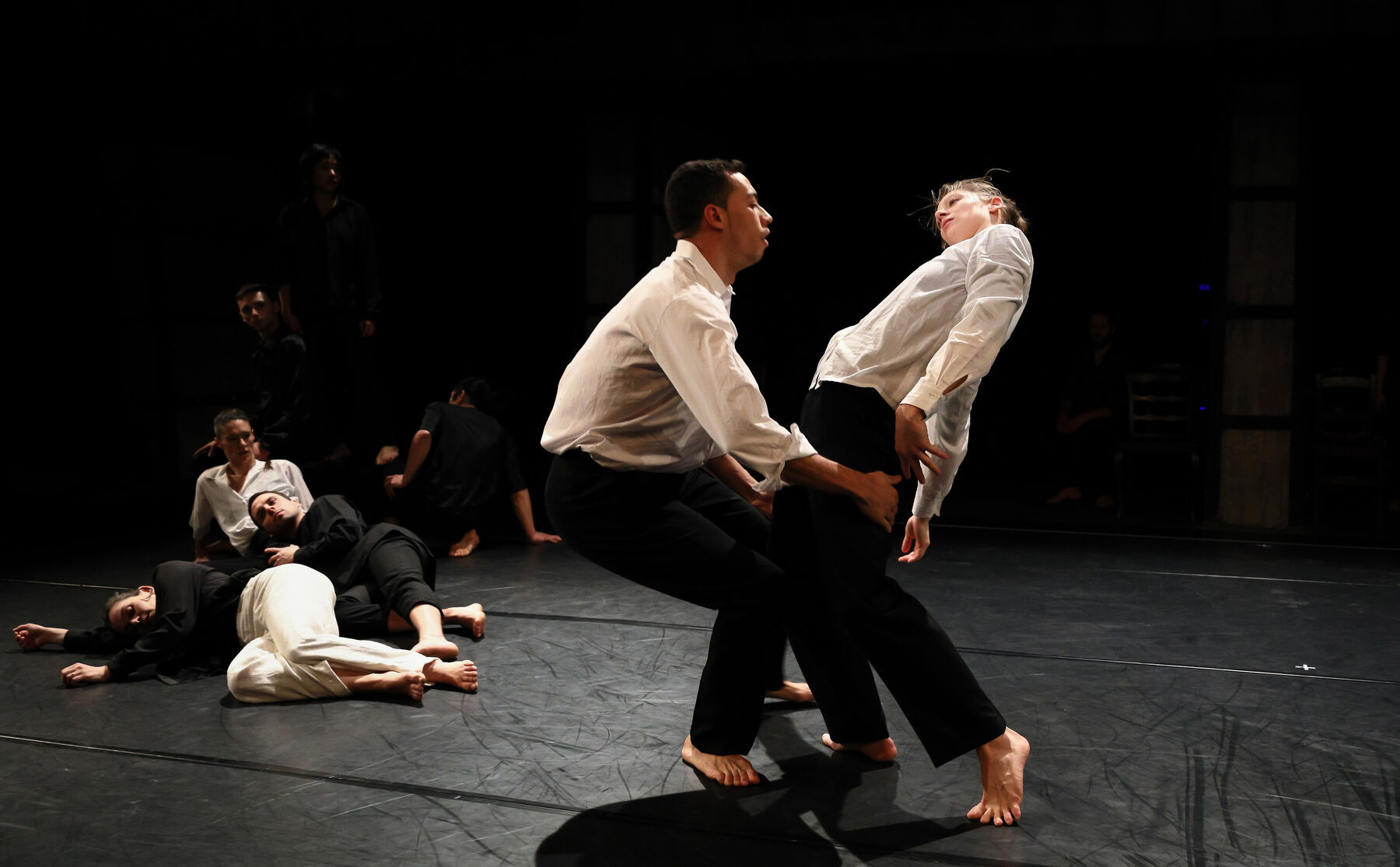 In the foreground, two dancers, dressed white, are dancing. In the back can be seen other dancers, lying on the floor.