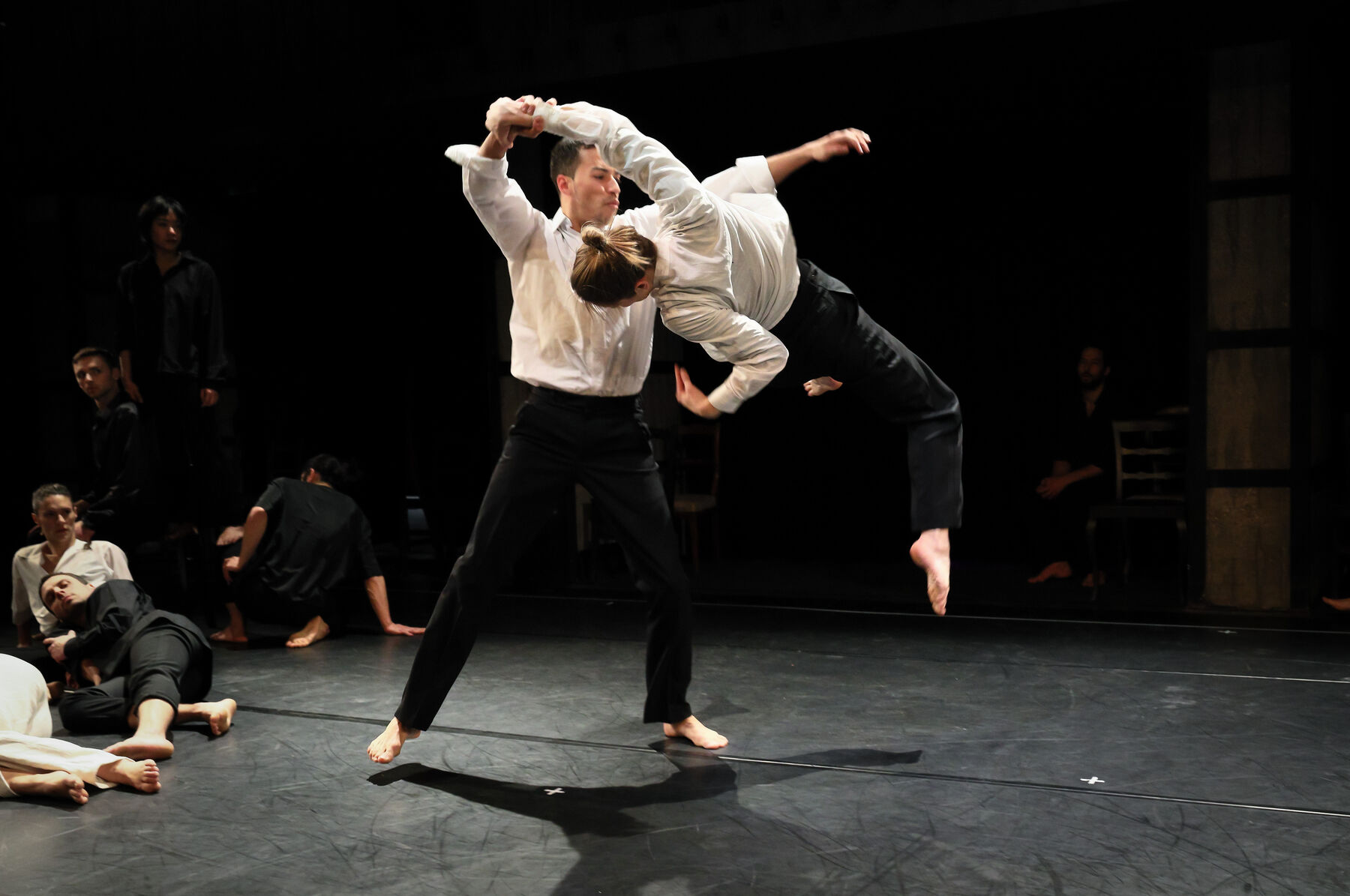 Two dancers, dressed white, are dancing together. In the background can be seen other dancers.