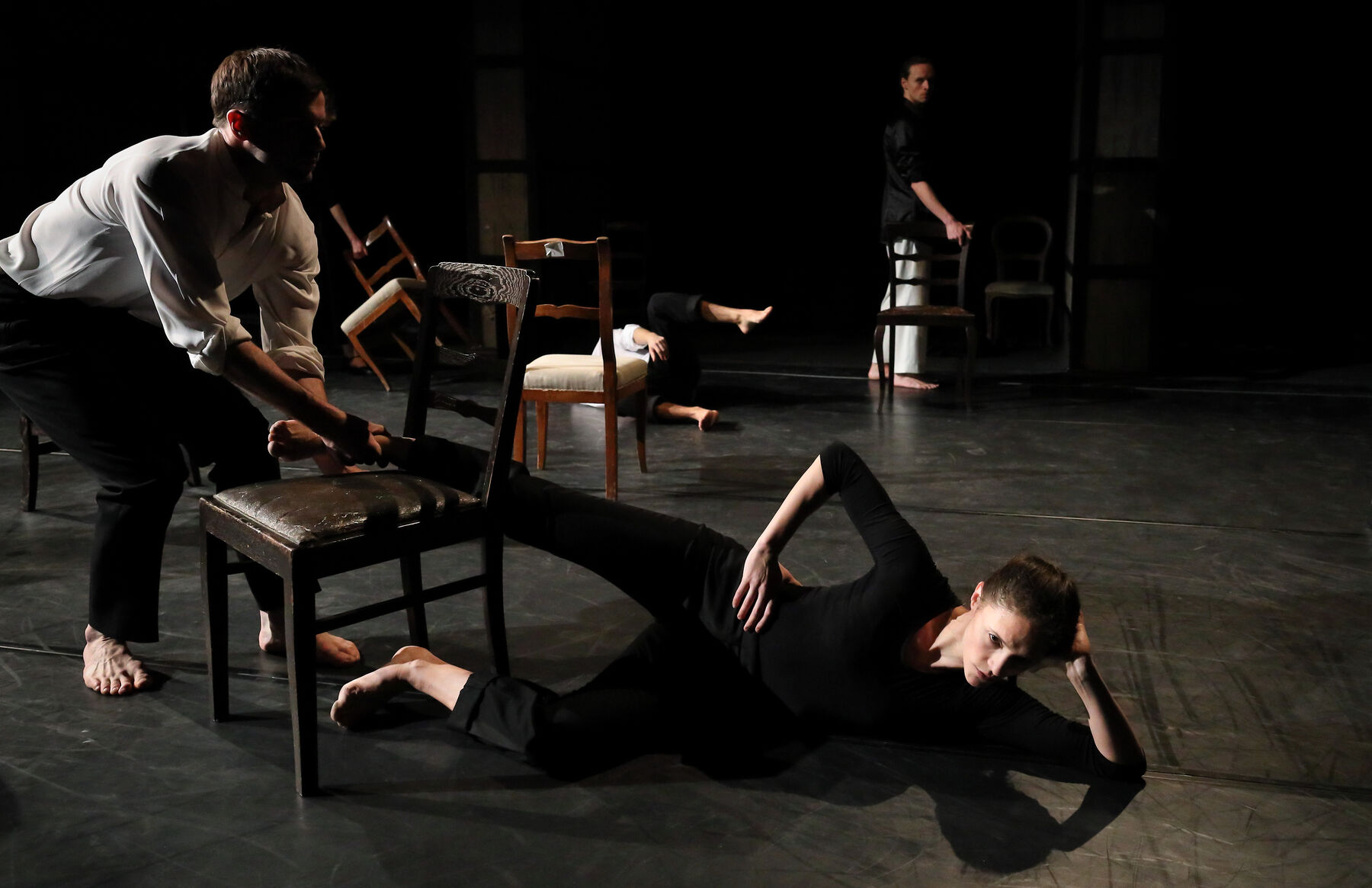 A dancer is lying o the floor. Chairs and two other dancers can be seen.