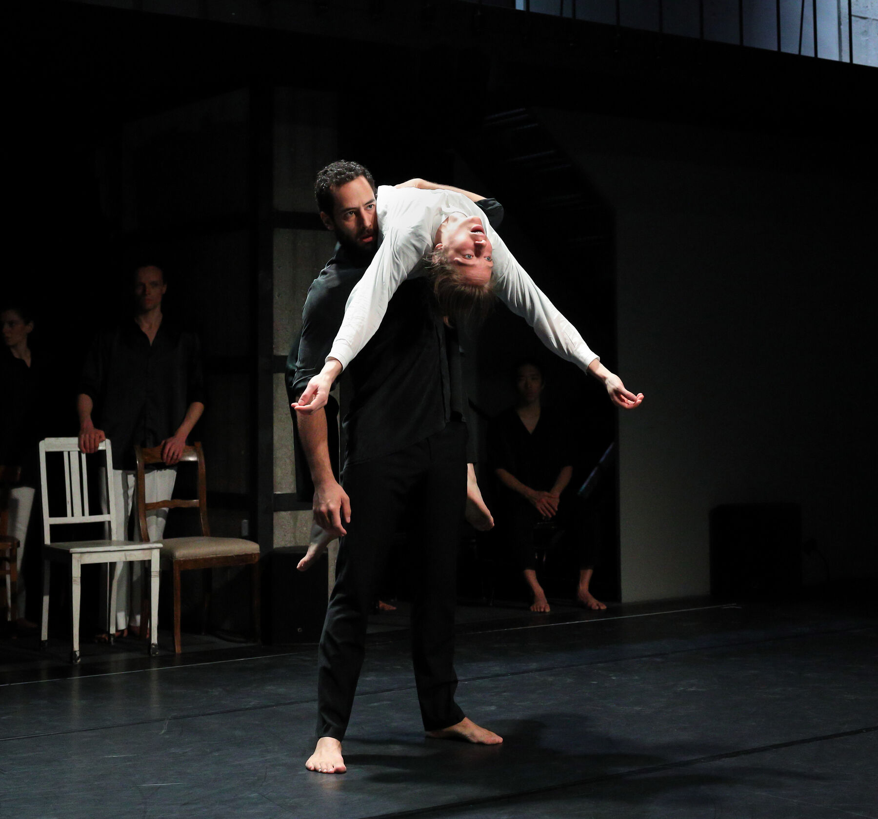 Tweo dancers are dancing in a dark room. In the background chairs and other dancers can be seen.
