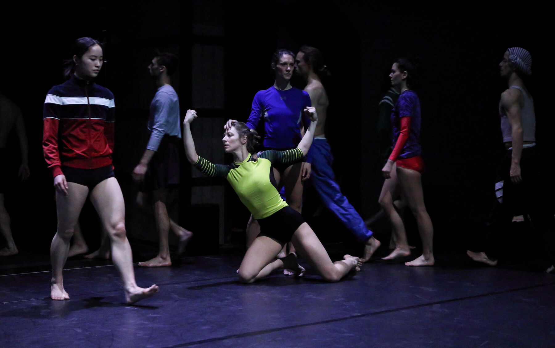 Dancers are walking in a dark room. In the middle another dancer is kneeling.