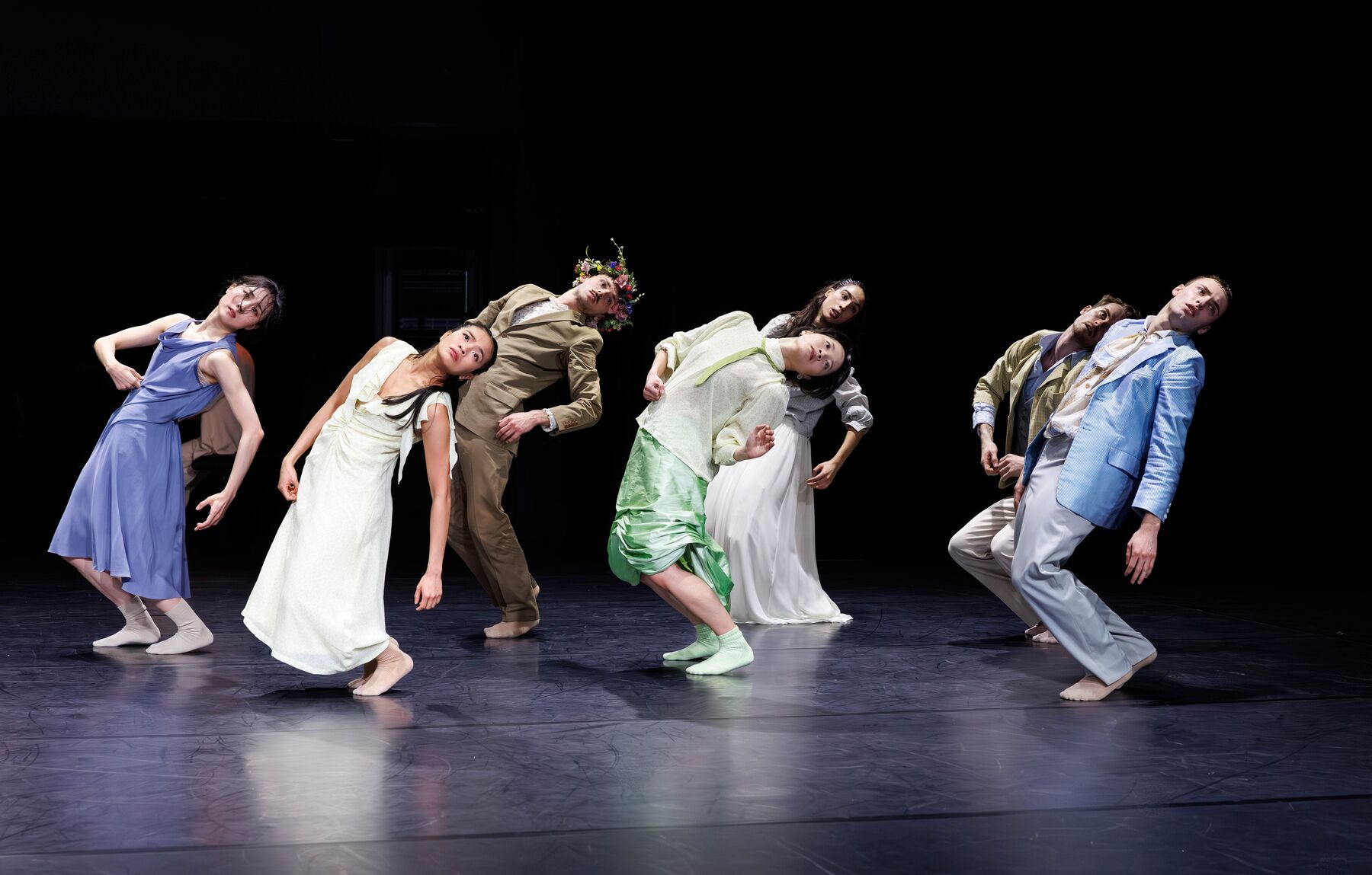 Seven dancers in bright, colorful costumes stand in two staggered rows. All of them are slightly bent at the knees and leaning back.