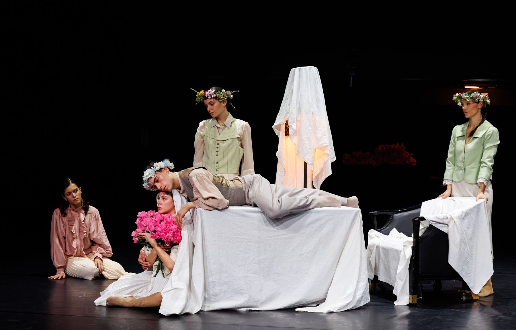 A table, a chair, and a lamp stand covered with white cloths in the middle. To the right of the chair stands a dancer wearing a flower wreath. In the center, a dancer wearing a flower wreath stands behind the table, while a dancer lies on the table, resting his head on the head of a dancer sitting on the floor. She is holding a bouquet of pink flowers in her hand. To the right of the table sits another dancer.