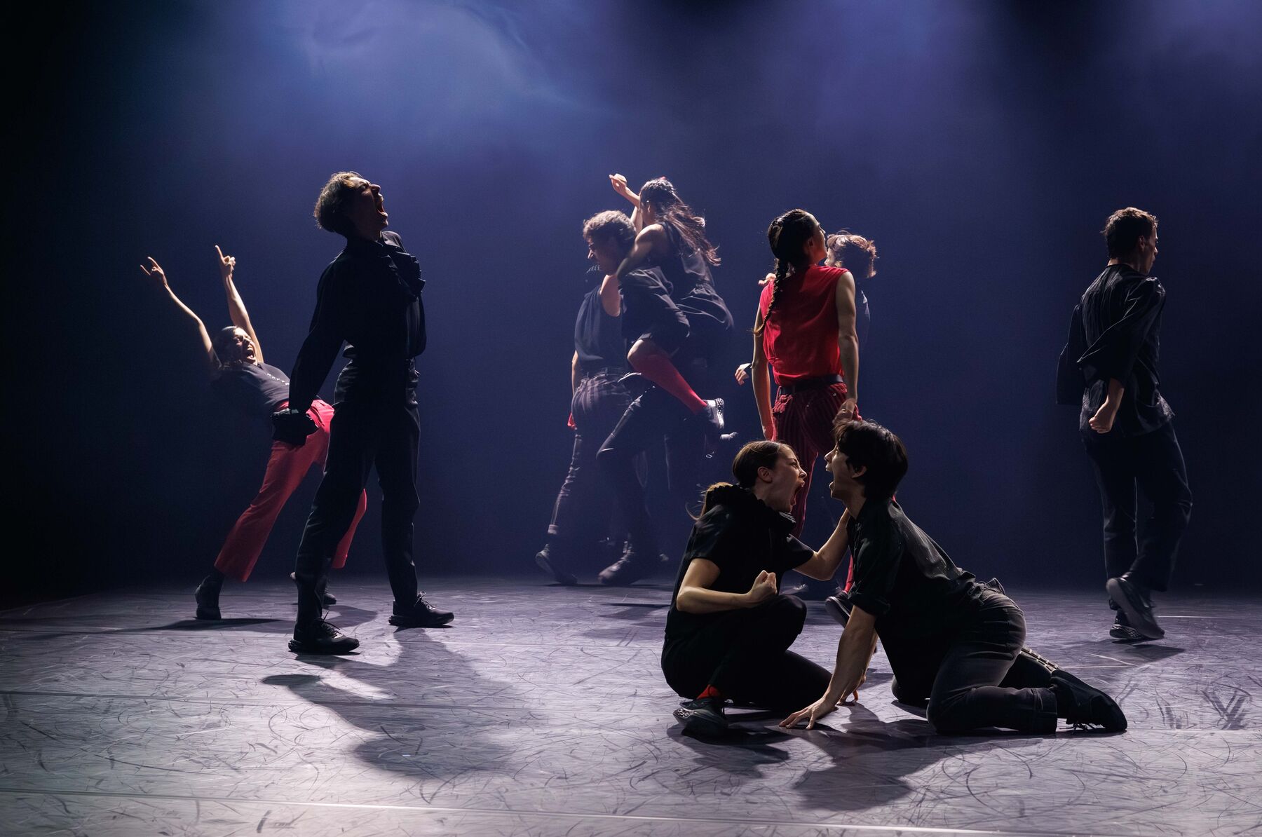 In a dimly lit room, there are several dancers dressed in red and black. In the foreground, two dancers are kneeling on the floor and shouting at each other. In the background, eight dancers can be seen shouting in various poses.
