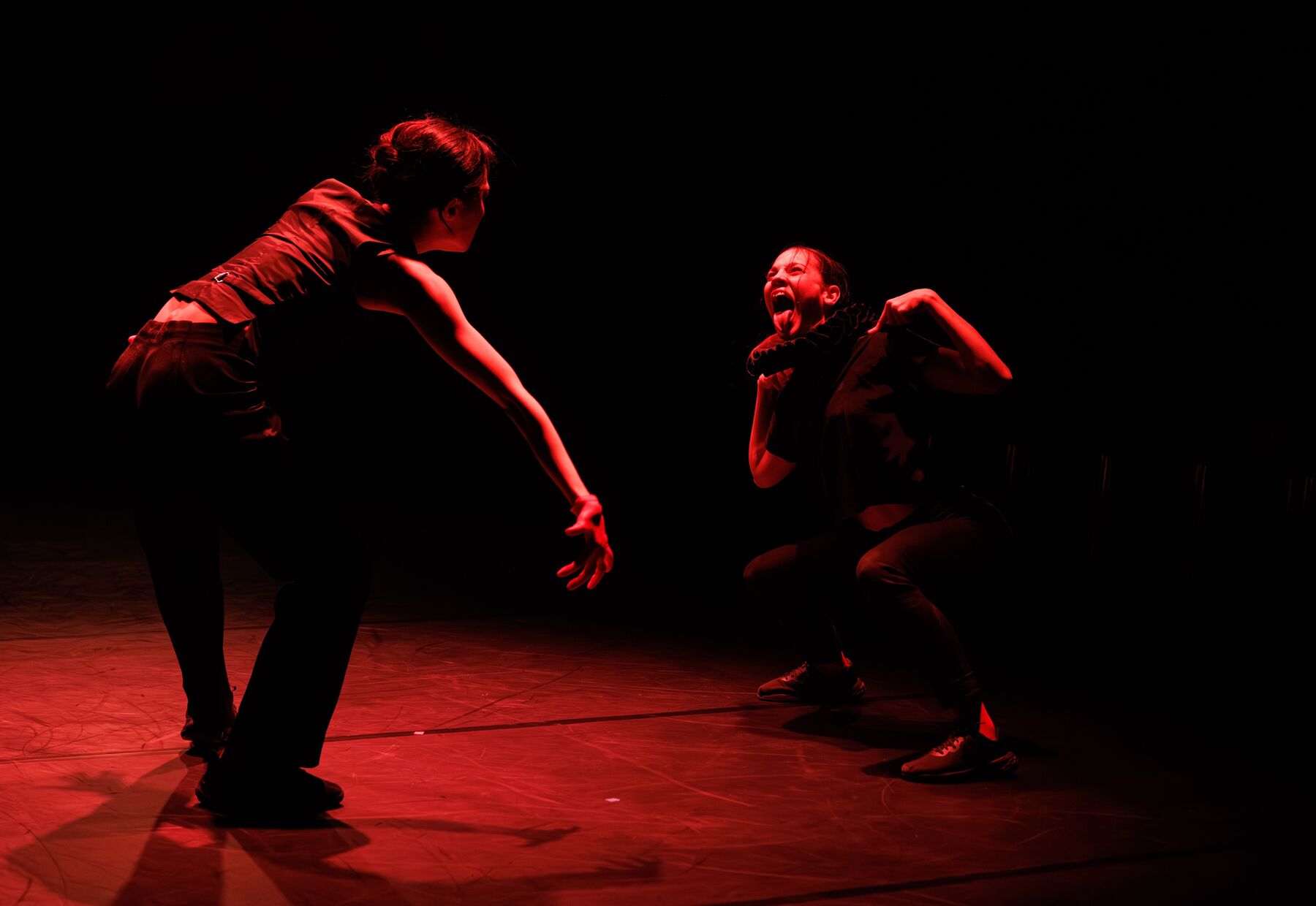 Two dancers in everyday clothes stand in a room lit by red light. The dancer on the left is seen from behind. She leans forward and stretches out her right arm. The dancer on the right is crouching, has her hands on her shoulders, and sticks out her tongue.