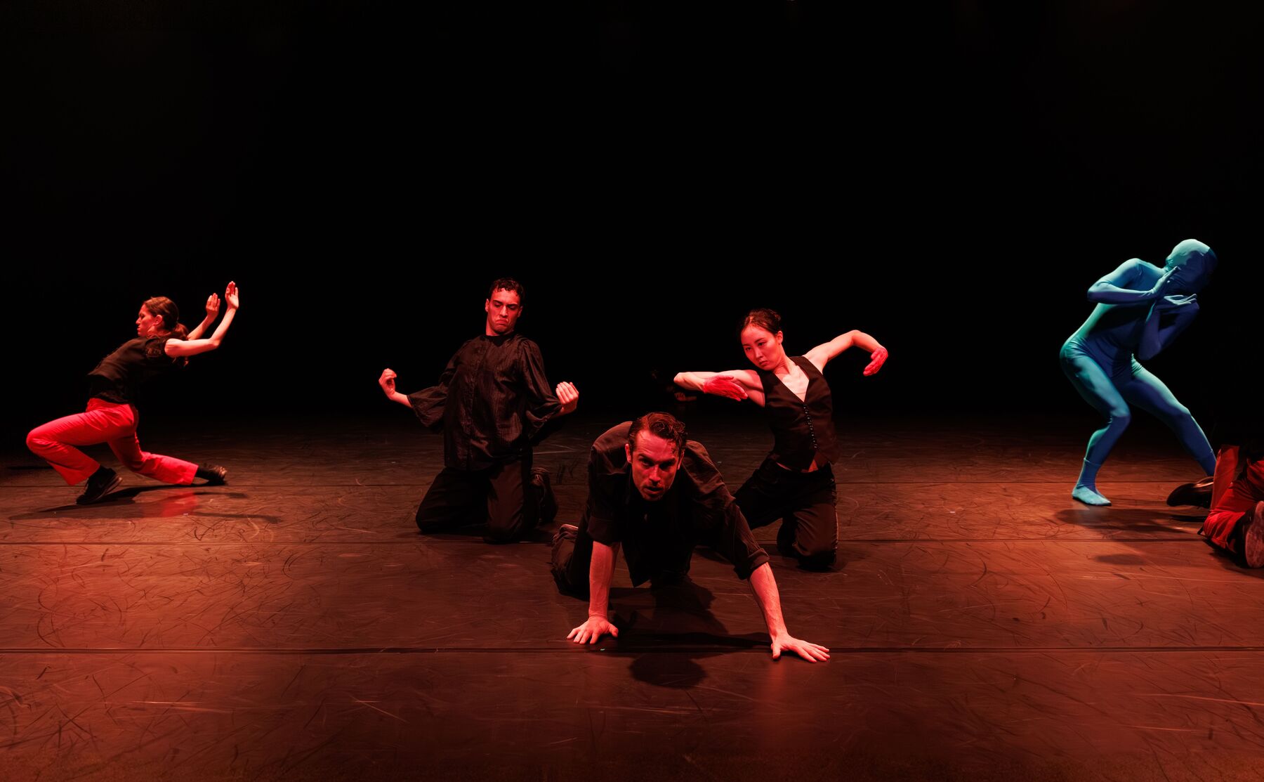 Five dancers dressed in black are in a room lit by dark red light. In the middle are three dancers who are on their knees and moving. The one in the middle is crawling toward the camera on all fours. In the background on the left is another dancer on her knees. On the right is a slightly bent dancer in a light blue full-body suit.