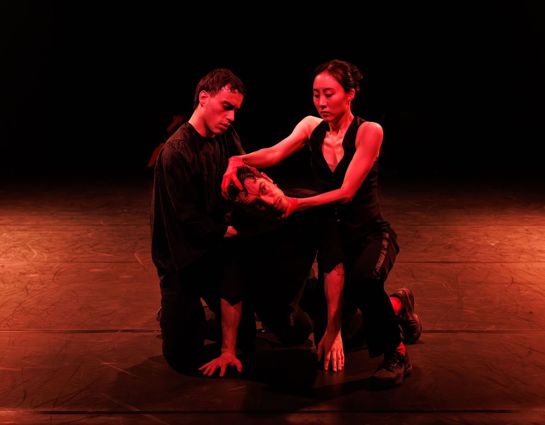 Three dancers kneel in a room lit by red light. The two on the outside hold the one in the middle and try to turn his head.