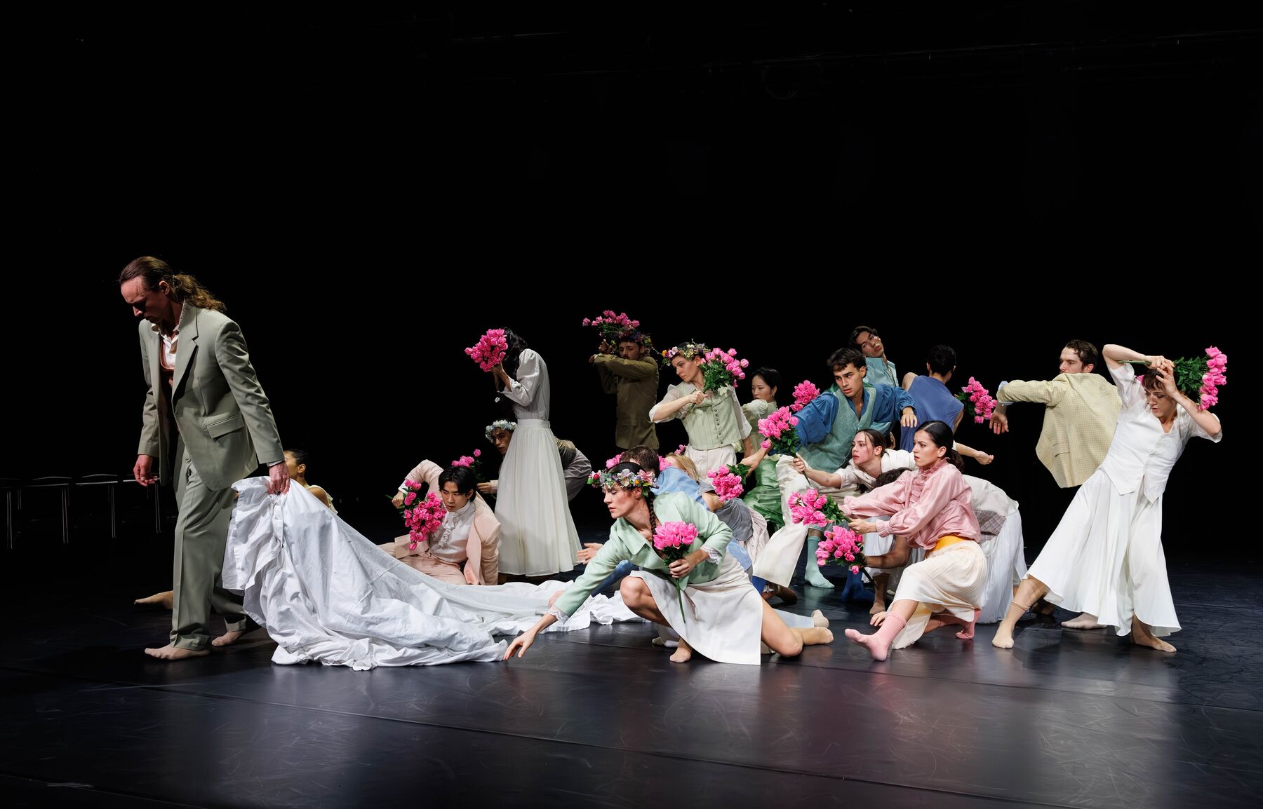 Many dancers wearing bright, colorful clothes, flower wreaths, and bouquets move around a white cloth, which is pulled away by a man in a light green suit.