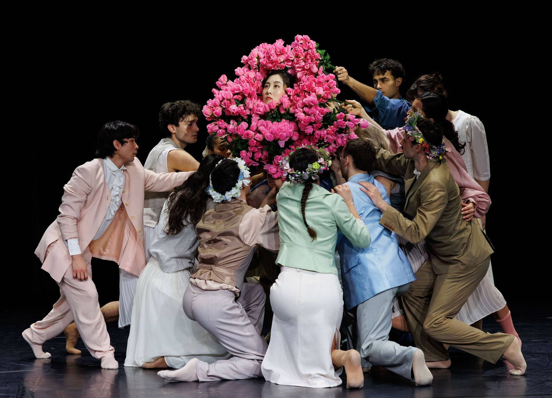 Many dancers wearing bright, colorful dresses and flower wreaths stand close together, holding pink bouquets in a circle around the face of a female dancer.