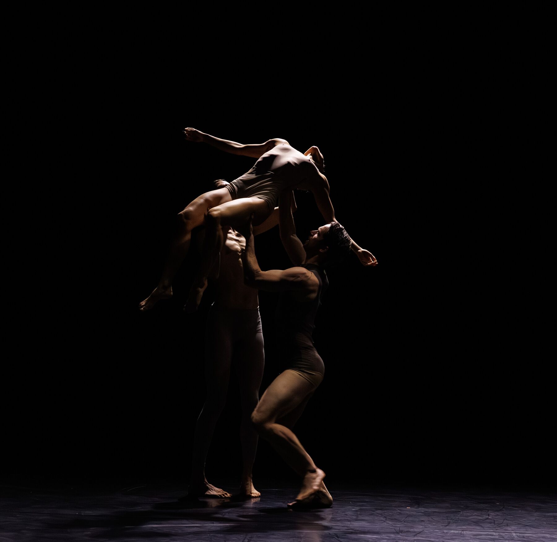 Three dancers in gray dancewear are standing in a dark room. Two male dancers are holding a female dancer aloft in a reclining position.