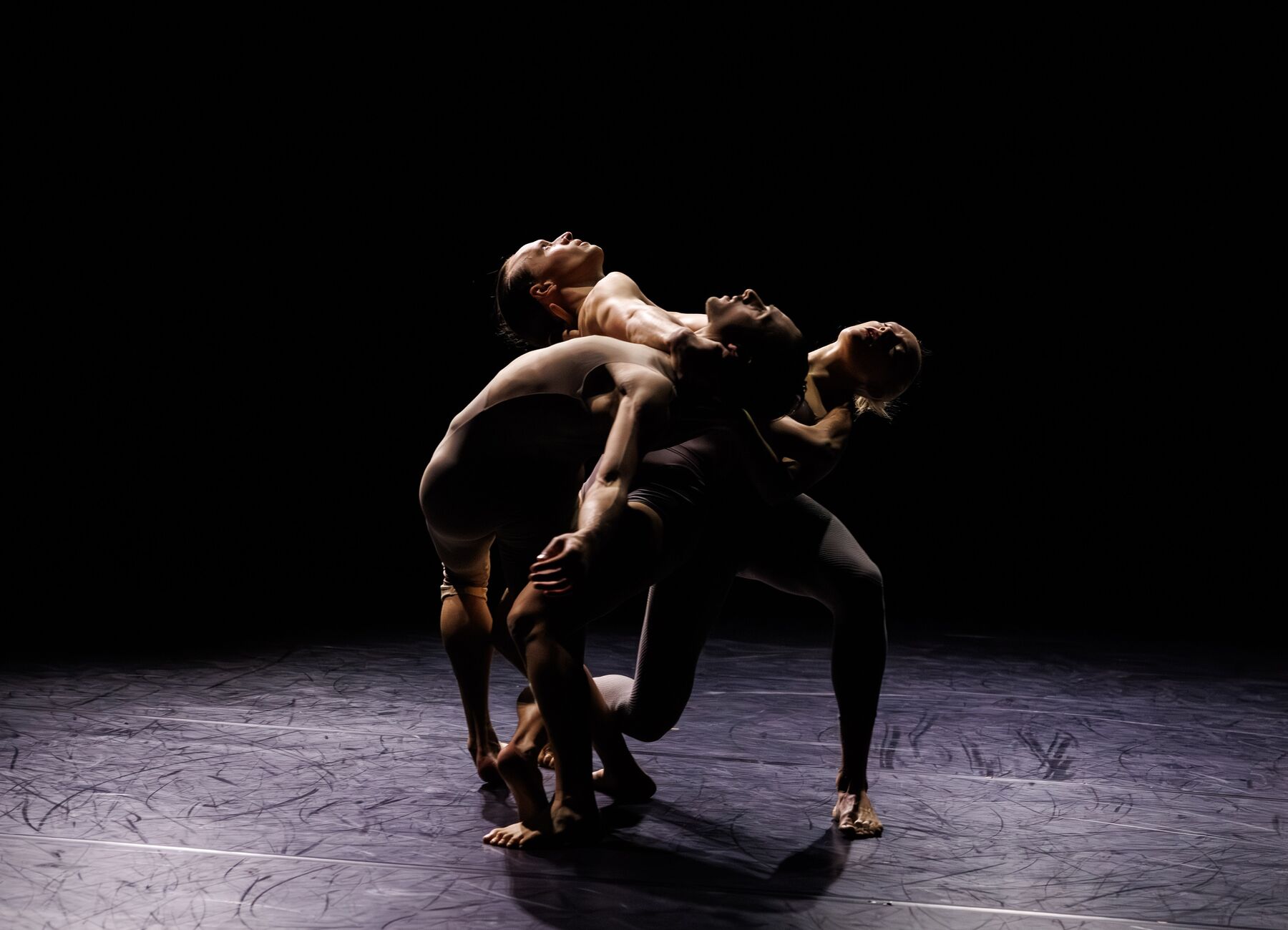 Three dancers in gray leotards are standing in a black room. The three dancers are standing close together and leaning against one another.