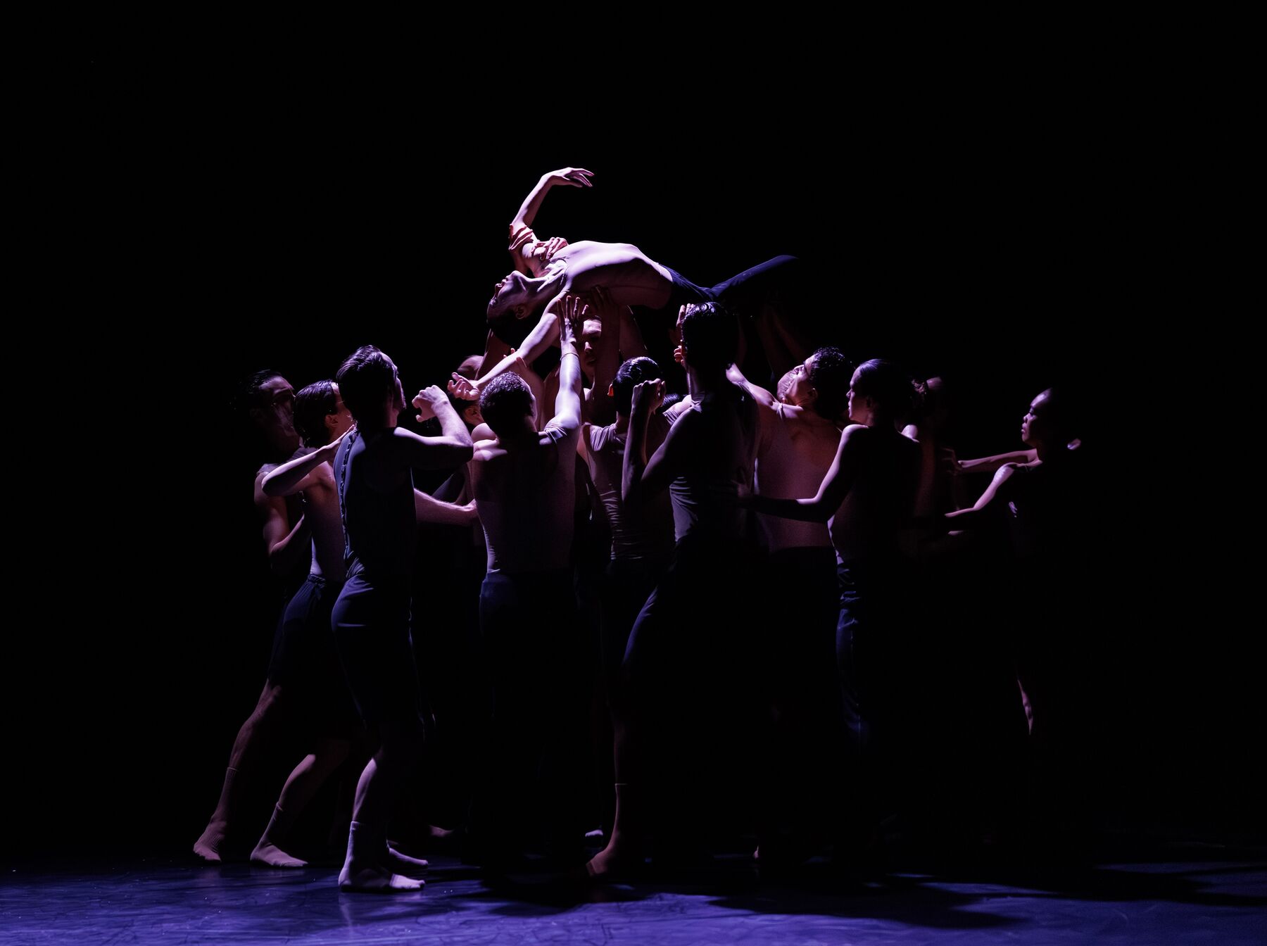 A group of dancers in dark, partly transparent clothing stands together in a crowded circle. A female dancer is lifted up by the group in the middle of the circle, lying on the back. The spotlight is on the dancer lifted up in the air.