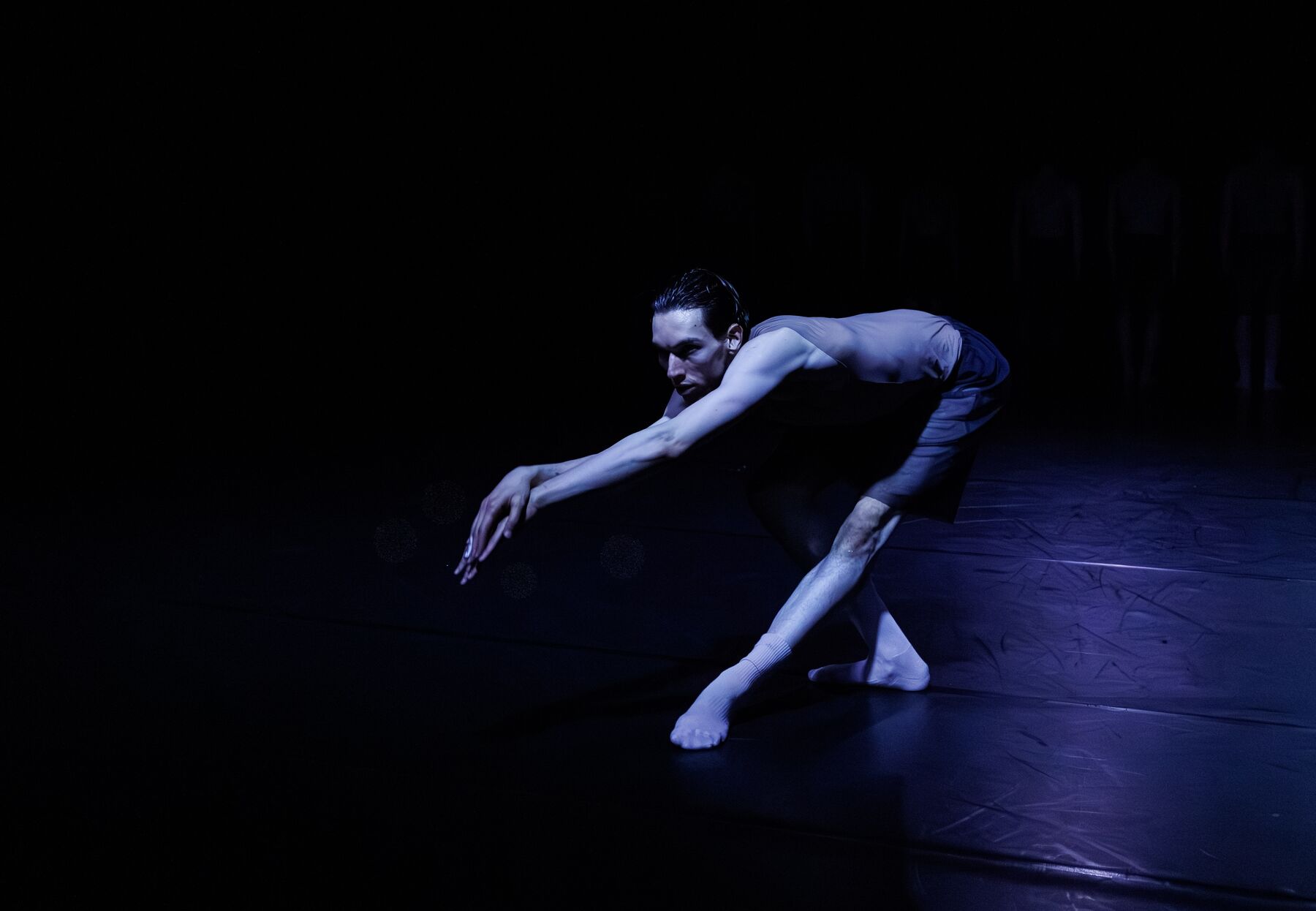 A dancer in dark, partly transparent clothes leans forward in a pose. One leg is stretched while the other is bent and the arms are stretched towards the front.