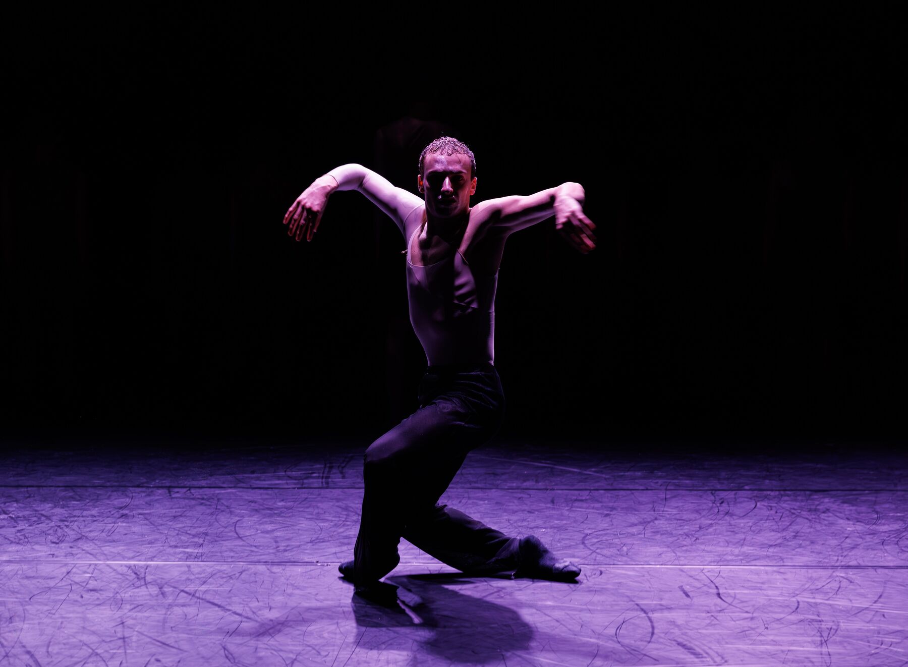 A dancer in dark, grey clothing is in motion. His legs are bent and his arms are slightly raised to the sides.