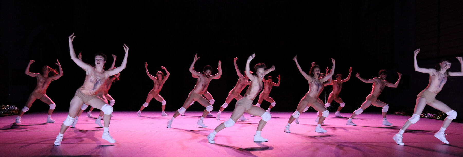Eine gruppe von Tänzern tanzt in einem Raum. Es ist dunkel und pinkes Licht scheint auf den Boden.