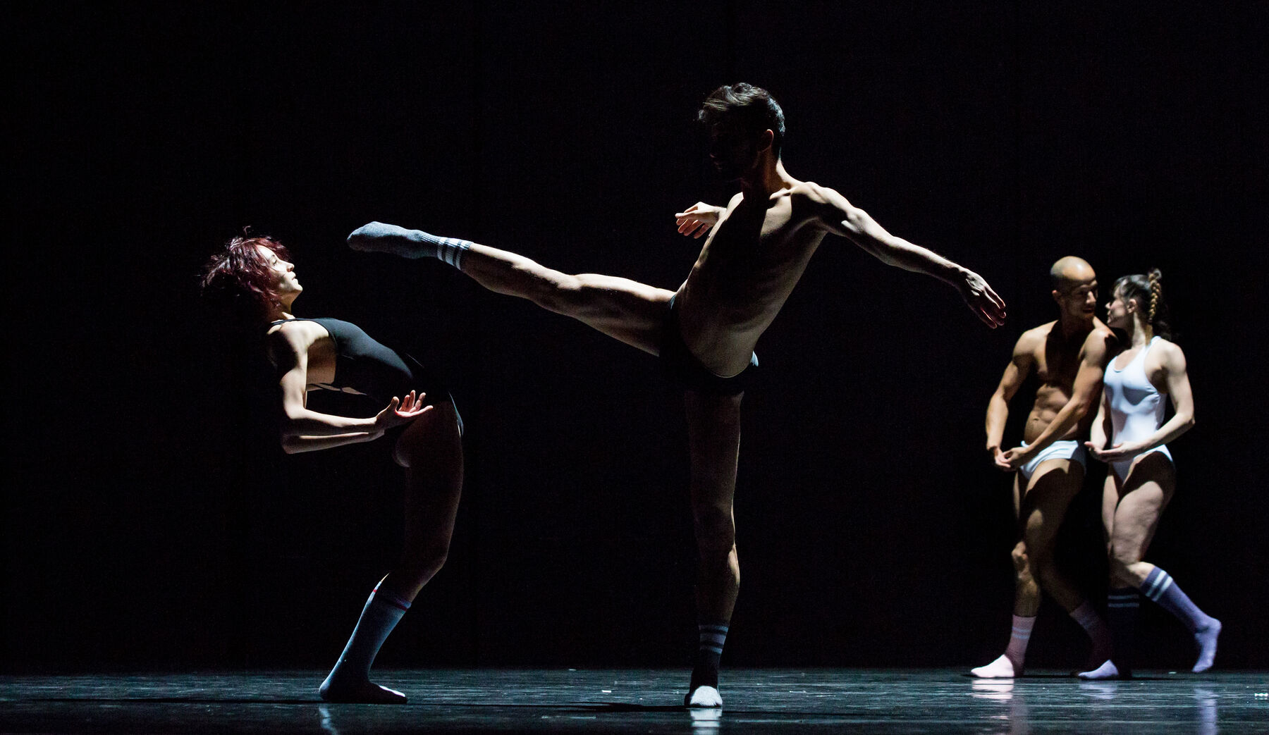 Ein Mann tanzt auf einem Bein, eine Frau daneben. Dahinter sieht man zwei weitere Personen.