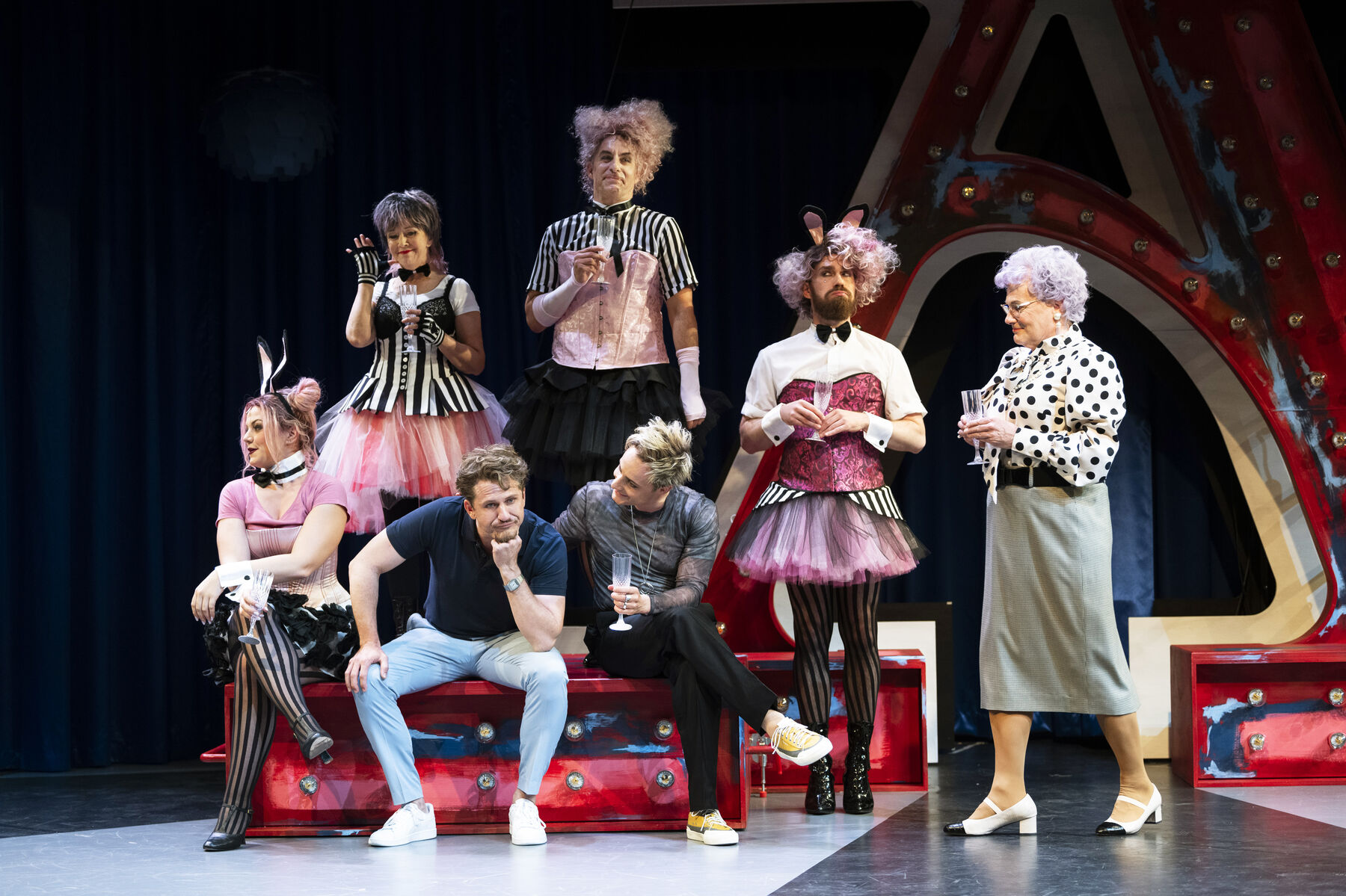 Two men are sitting on a red crate. One of them is holding a champagne glass. Behind them are four people in pink and black costumes with corsets and bunny ears. On the right of the picture is an old woman with a champagne glass in her hand. 