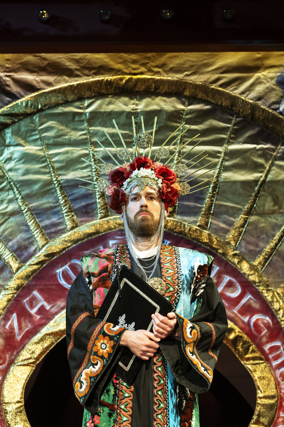A man in a kind of saint's costume with a saint's crown stands in front of a golden background and holds a book in his hands. 