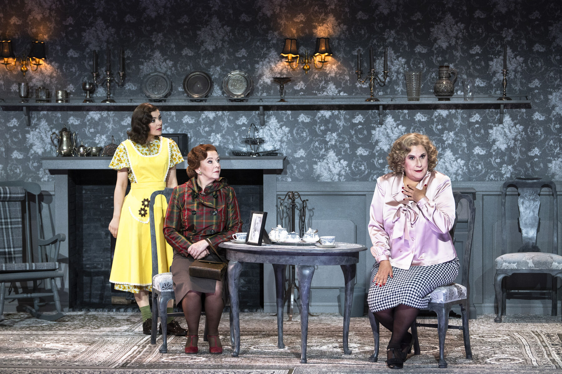 Miss Pinkerton and Miss Todd are sitting in Miss Todd's living room having tea. Miss Todd clutches her chest in shock. Laeticia stands beside them and looks at Miss Todd with concern. 