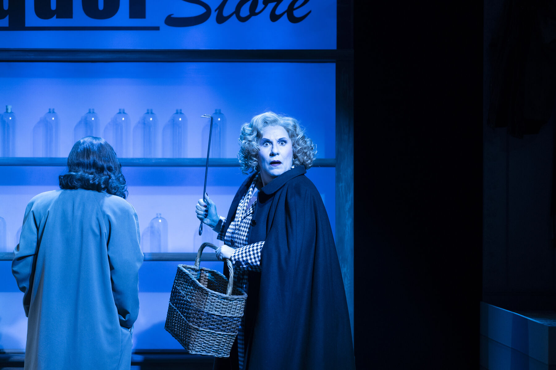Miss Todd looks frightened into the camera, holding a crowbar in one hand and a woven basket in the other. Behind her stands Leticia, with her back to the camera. The two are in a liquor store. The scene is lit with blue light. 