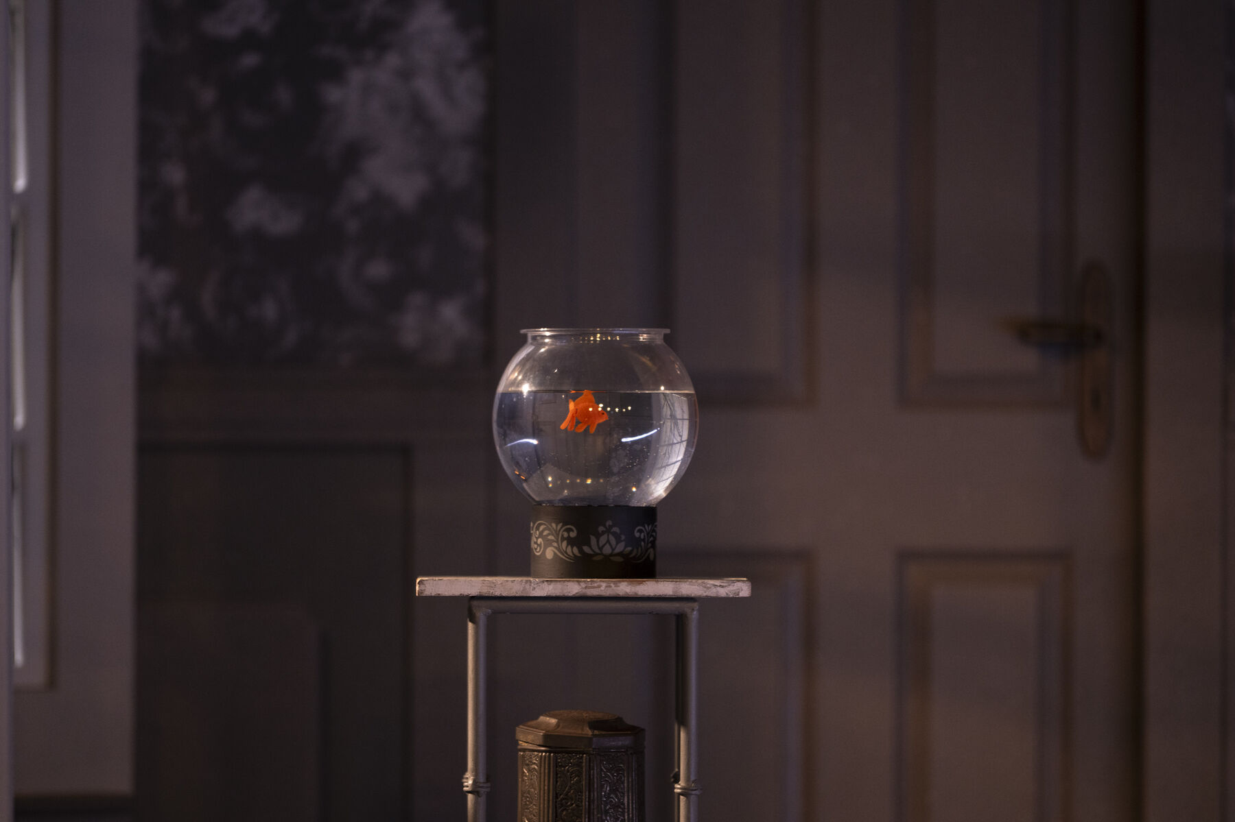 A goldfish bowl stands on a table, a goldfish swims in it.