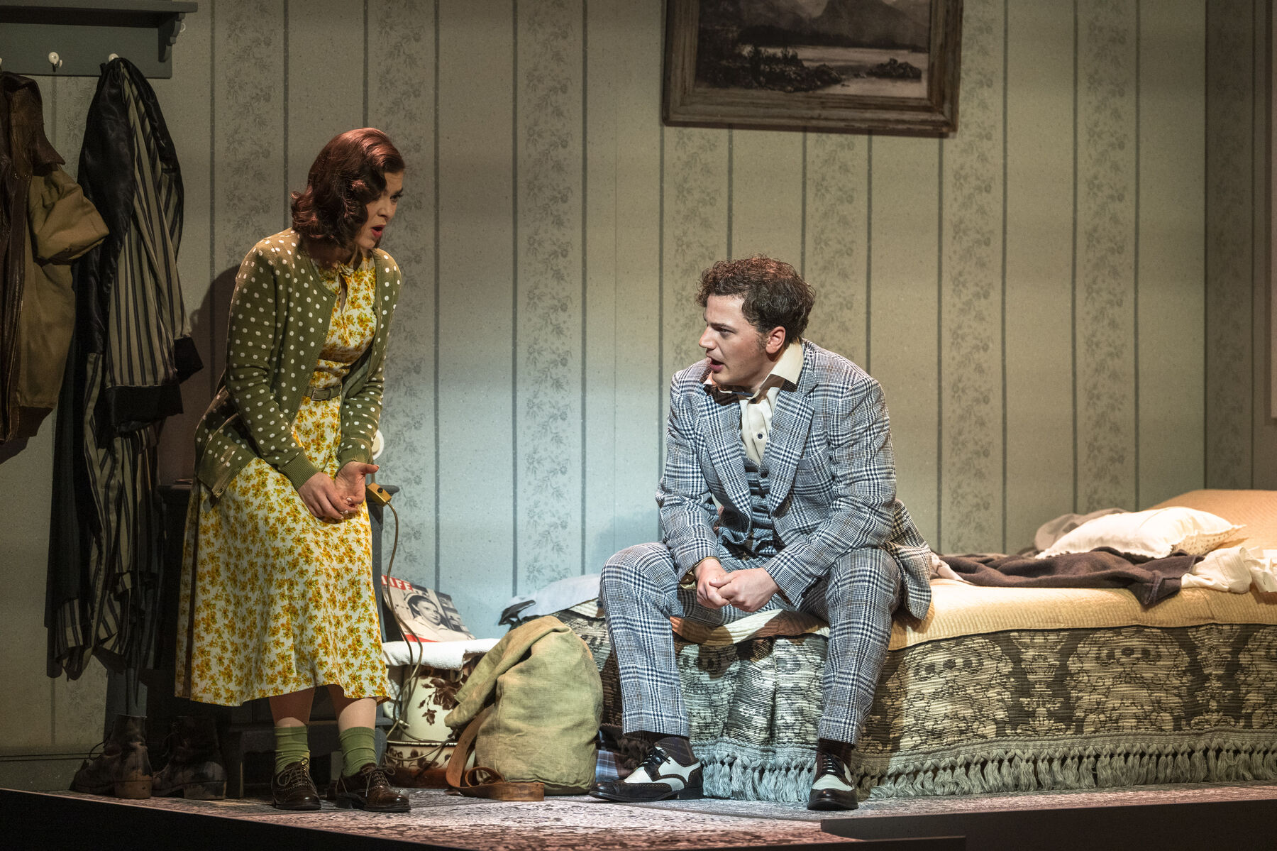 Bob is sitting on the bed talking to Laetitia, who is standing opposite him. She is distraught. 
