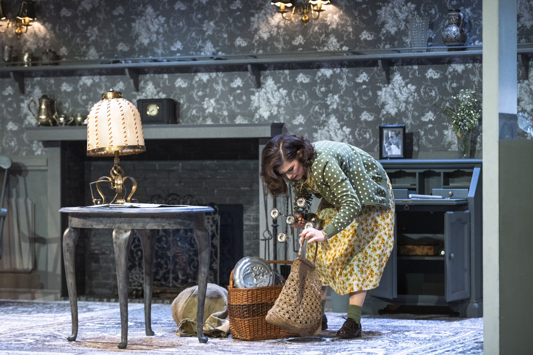 Laetitia is in Miss Todd's living room. In front of her lies Bob's rucksack and a large basket with silver cutlery. She bends over a bag into which she is hurriedly placing a silver candlestick. 