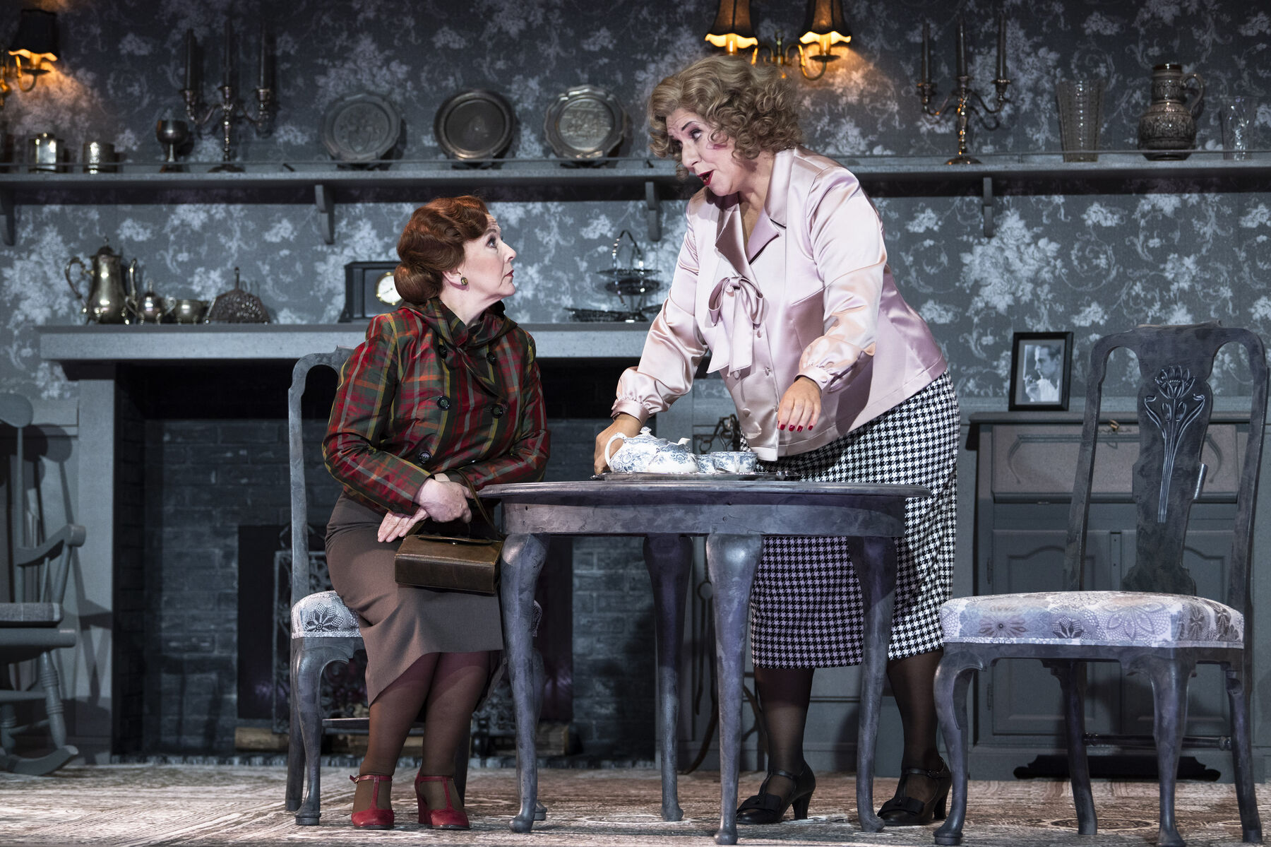 Miss Pinkerton is sitting at the table, Miss Todd is bringing in a tray with tea. The two look at each other. Miss Todd's living room is classically furnished and rather dark and cool.