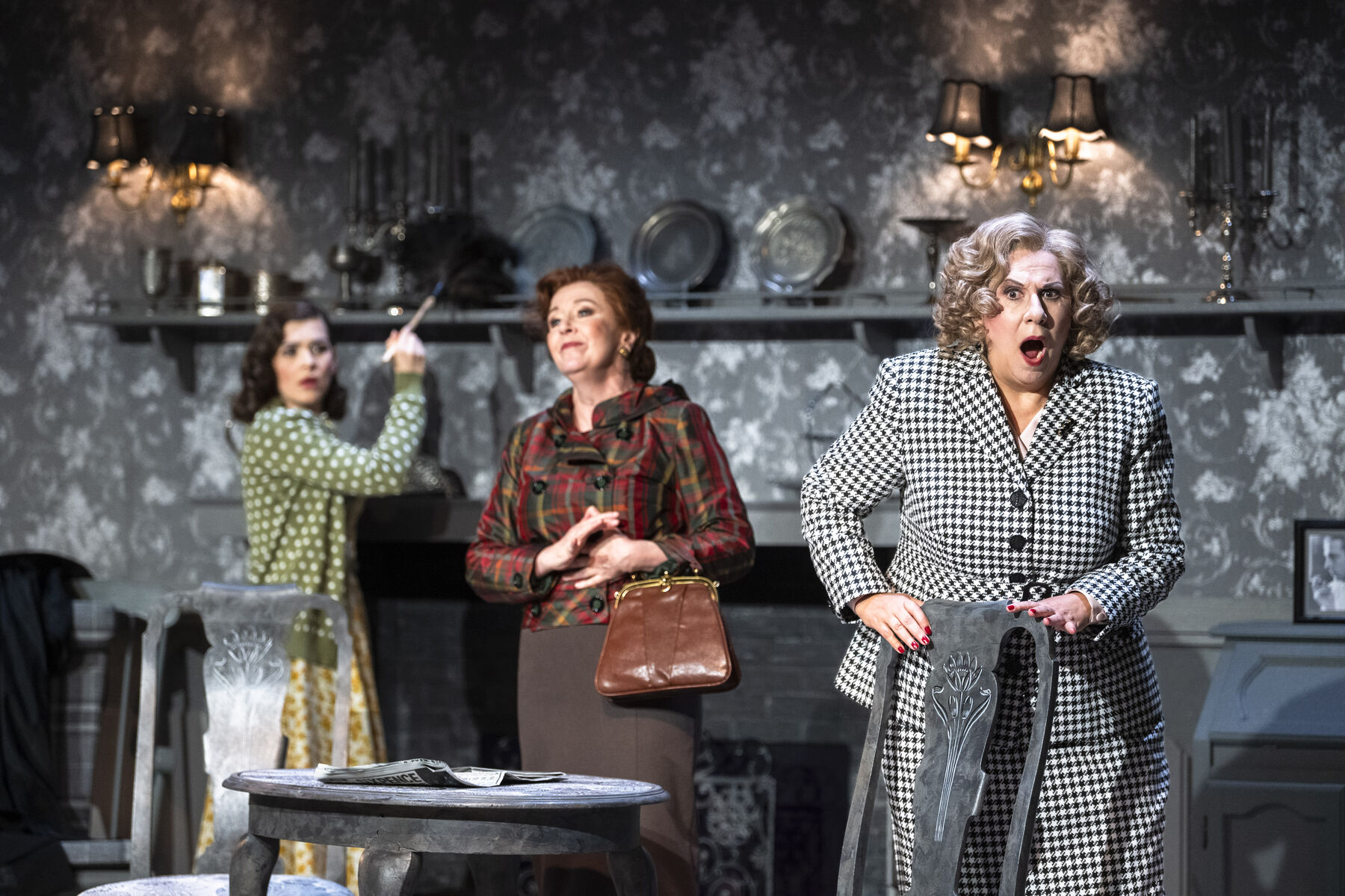 Miss Pinkerton is standing at a table with an open newspaper in front of her. She smiles contentedly. Miss Todd is very upset, clutching the back of a chair with her mouth agape. Laetitia is dusting a wall shelf and looks at Miss Todd in disbelief. 