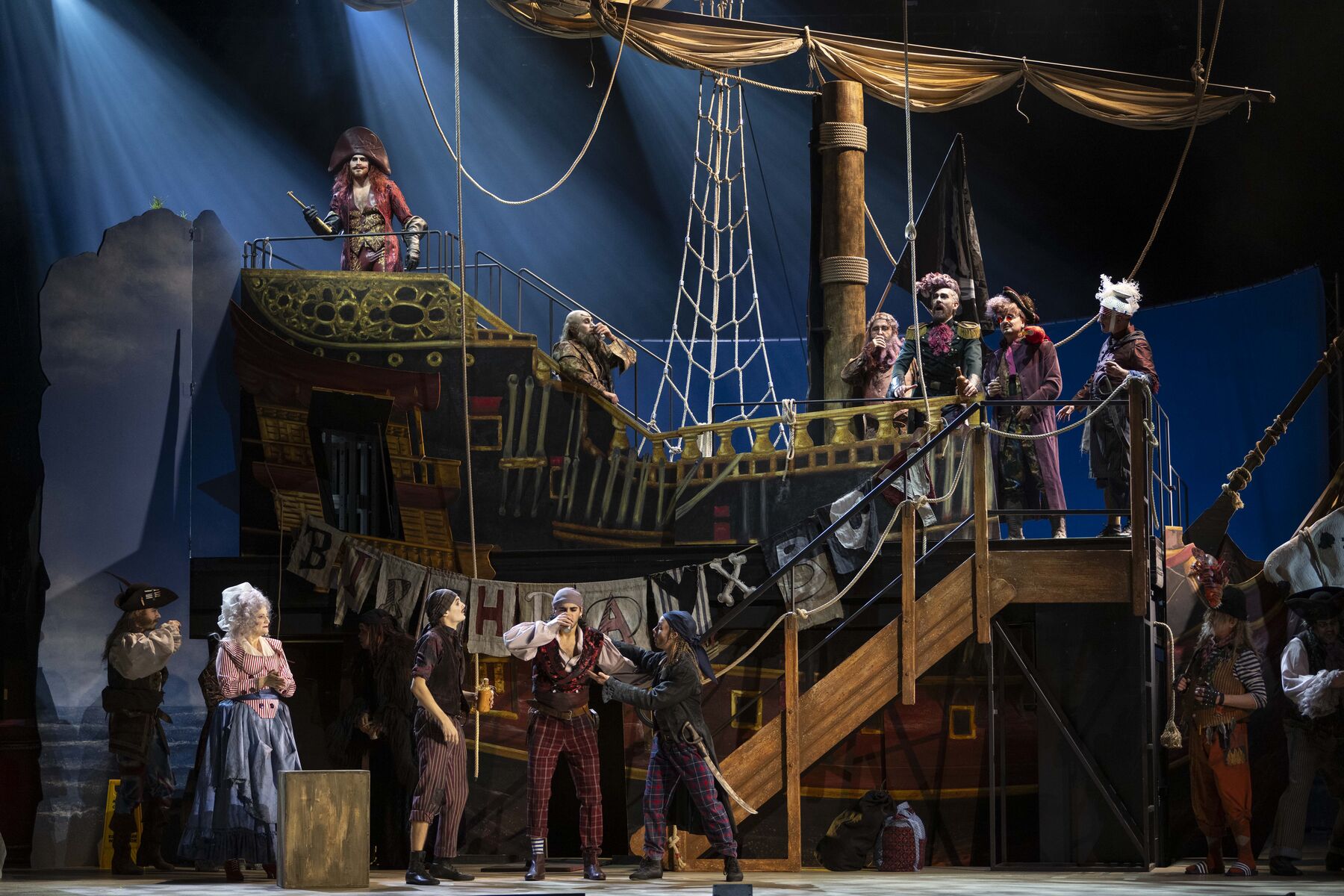A large pirate ship. A large garland is hung up with the words ‘BIRTHDAY BOY’. The pirate king stands at the highest point of the ship, holding a telescope in his hand. Below him stands a young pirate, Frederic, who is being asked to empty his cup. Several other pirates are scattered around the ship. 