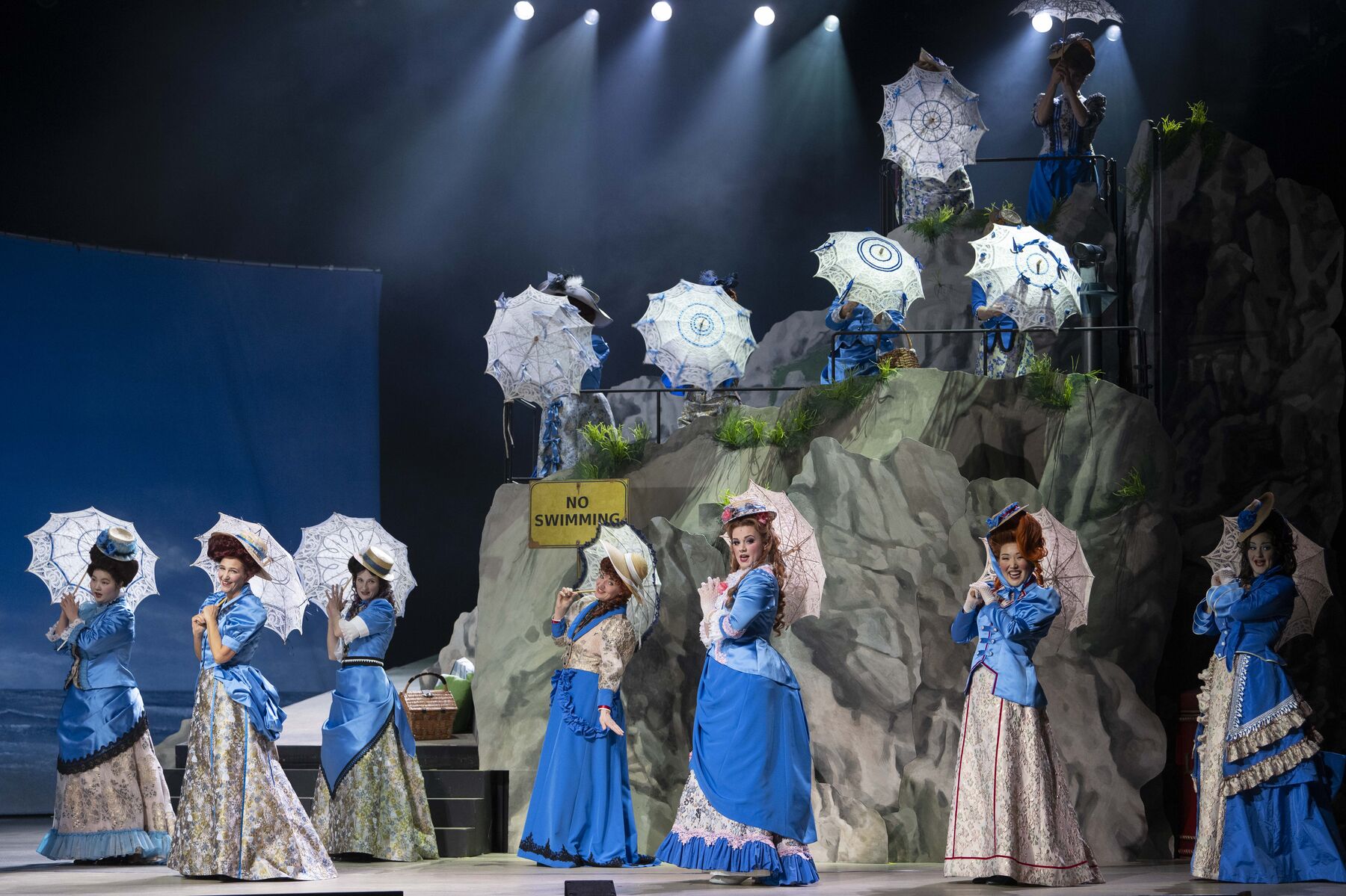 Twelve young ladies in blue dresses are standing on a beach with cliffs. They are all wearing hats and holding up lace parasols. A sign is attached to the rocks with the words ‘no swimming’.