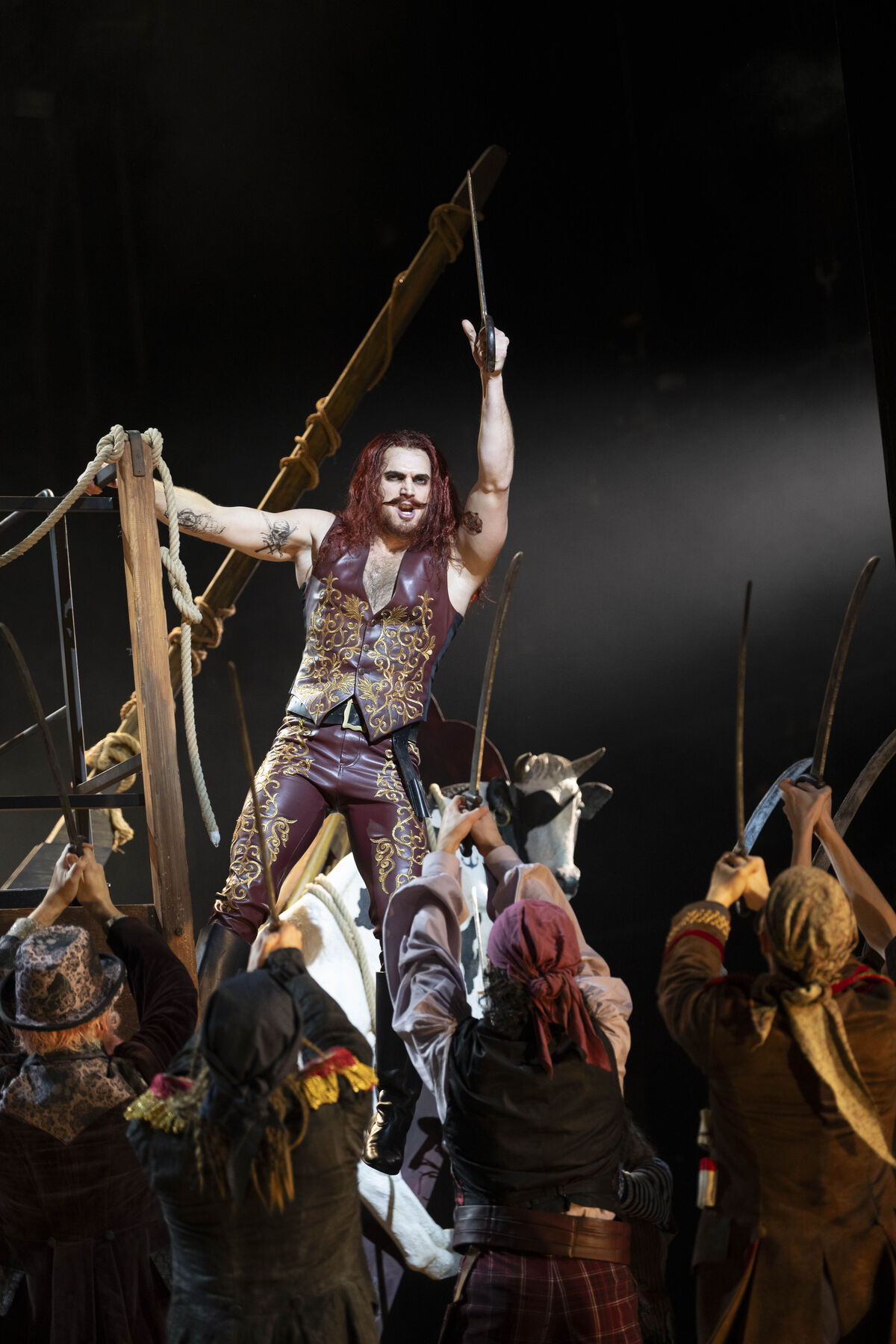 The pirate king holds onto a wooden scaffold. He raises his sabre. Some pirates look up at him and raise their sabres in agreement. 