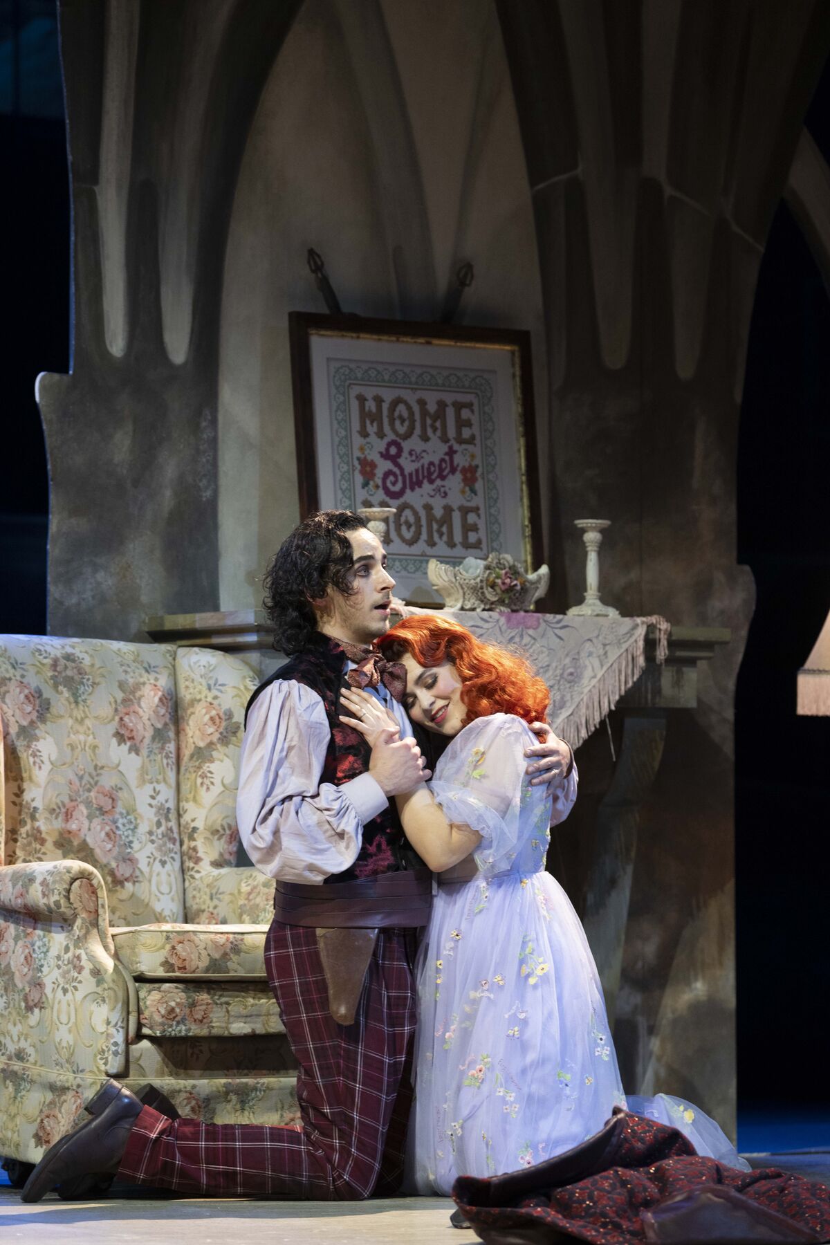 Mabel and Frederic are both kneeling upright on the floor in the Major General's house. She lovingly rests her head on his chest and closes her eyes. Frederic puts his arms around her. In the background there is an armchair and a fireplace. On the mantelpiece there is a picture frame with the words ‘home sweet home’. It is night-time.