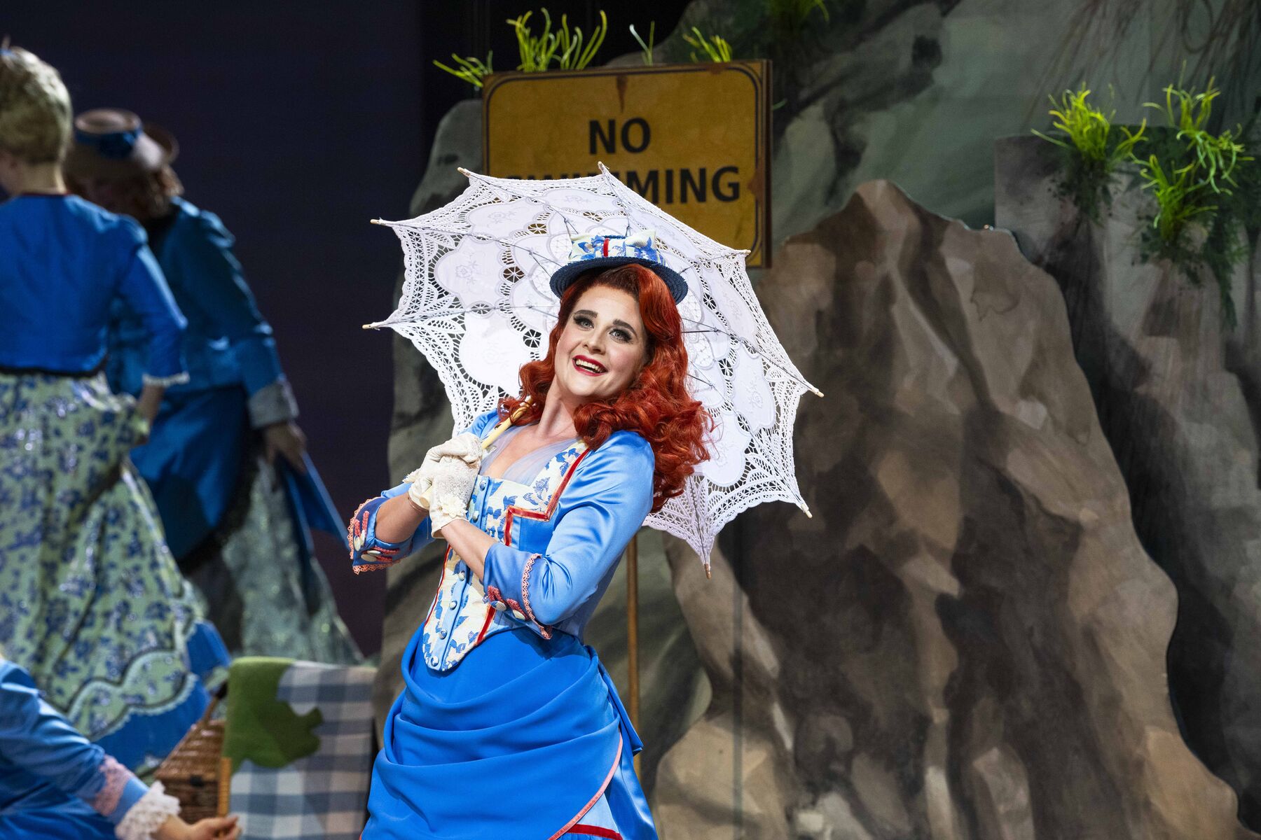 The focus of the picture is a young woman. Mabel poses smiling with her lace parasol. She has bright red hair and wears a light blue dress. In the background are rocks and a few other wards in similar clothing.