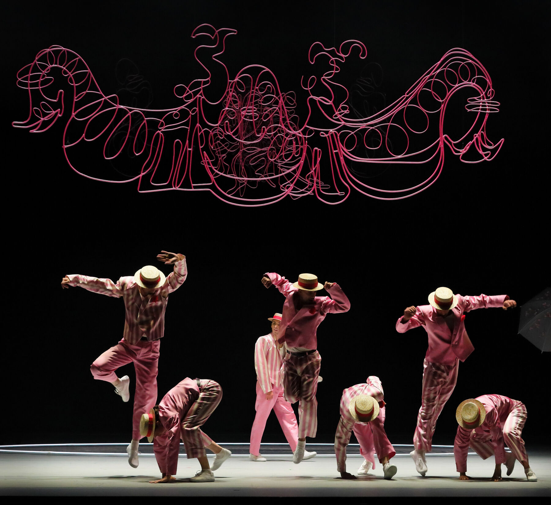 Sieben Tänzer in rosa Anzügen tanzen in zwei reihen. Sie tragen Strohhüte mit roten Hutbändern. Über ihnen hängt ein rotes verschlungenes Drahtkonstrukt. Der Rest der Bühne ist schwarz.