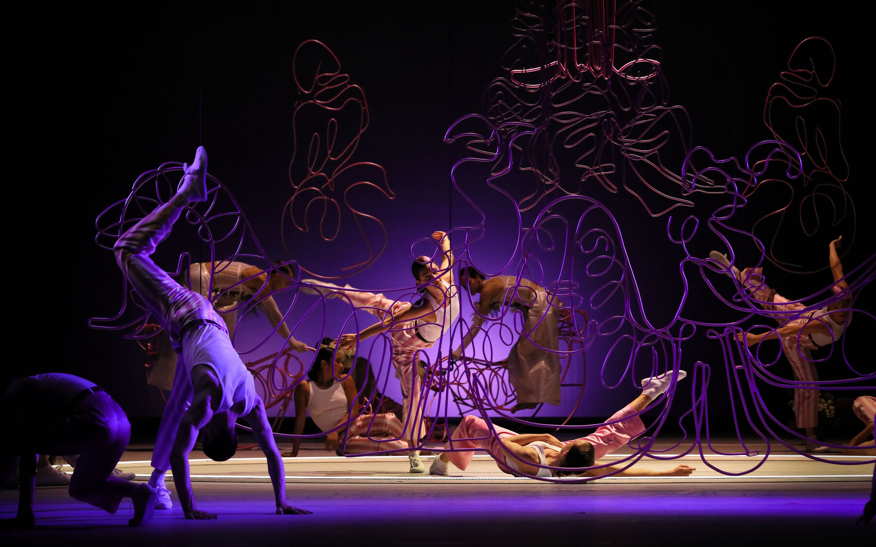 Im Fokus steht eine Tänzerin in gestreckter Haltung mit einem erhobenen Bein und ausgestreckten Armen. Um sie herum befinden sich weitere Tänzer und Tänzerinnen und verschiedene geschwungene Drahtinstallationen in rosa, pink oder lila. In der Szene herrscht viel Dynamik.